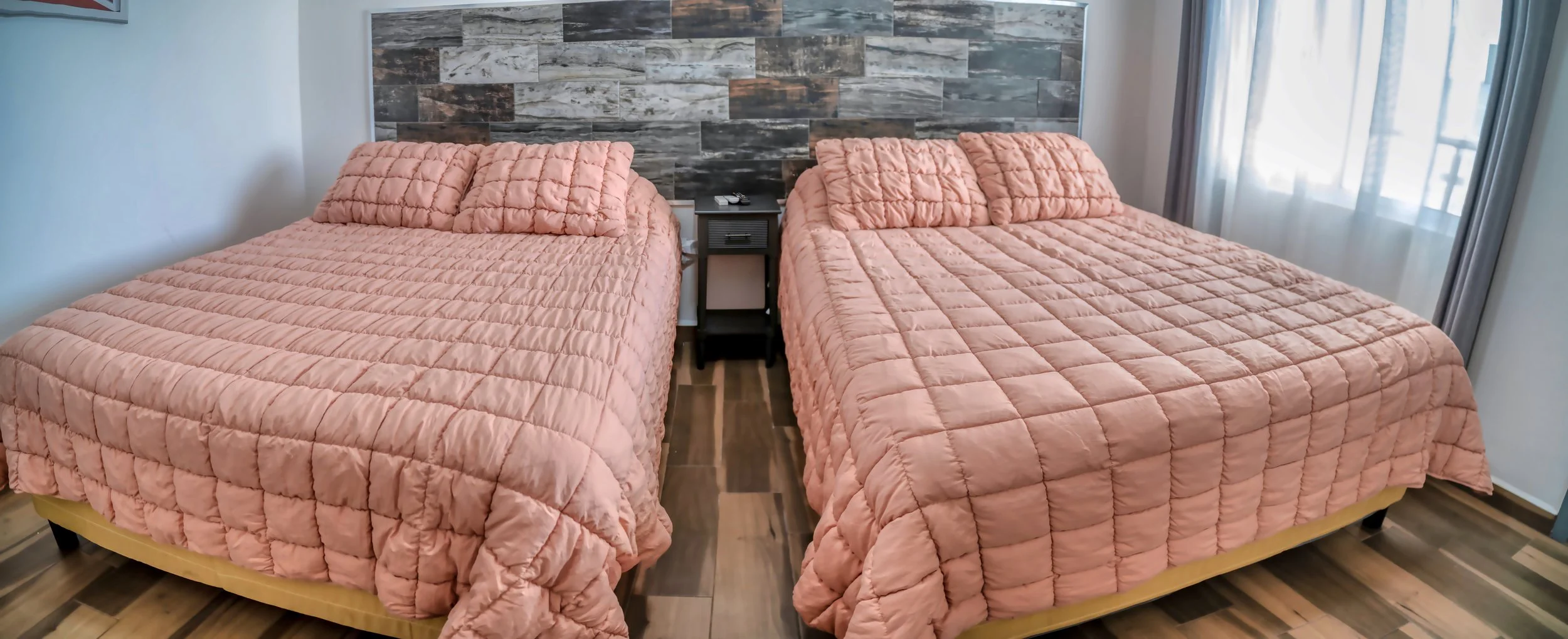 Dos camas dobles con cubrecamas rosados y almohadas a juego, una mesa de noche entre ellas, en una habitación con paredes blancas y una pared de acento de piedra, y cortinas blancas y grises en la ventana.