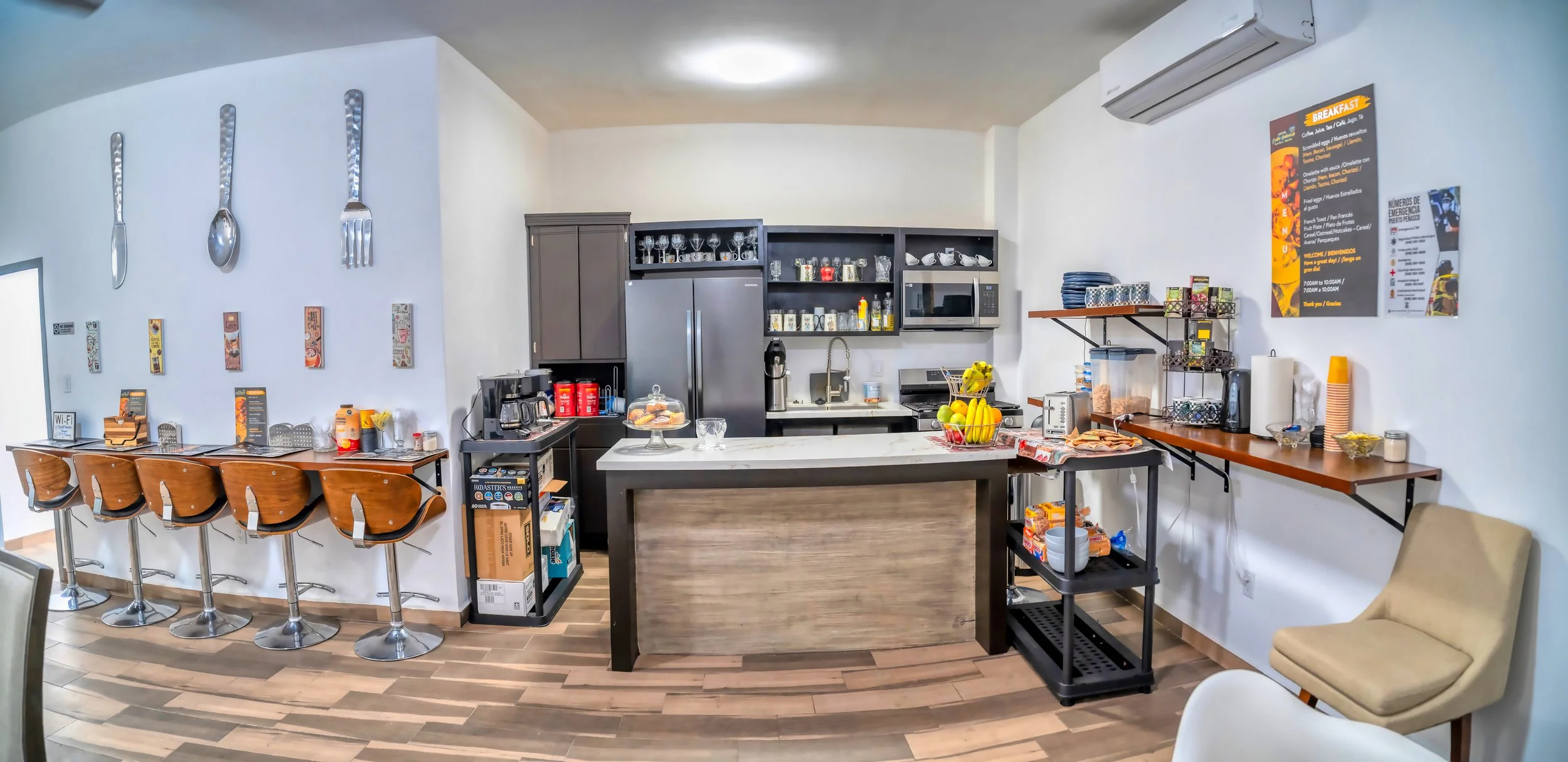 Cafetería moderna con barra de madera, sillas de bar, estanterías con tazas y platos, área de preparación de alimentos con frutas y pan, electrodomésticos como microondas y cafetera, y cartel informativo en la pared.
