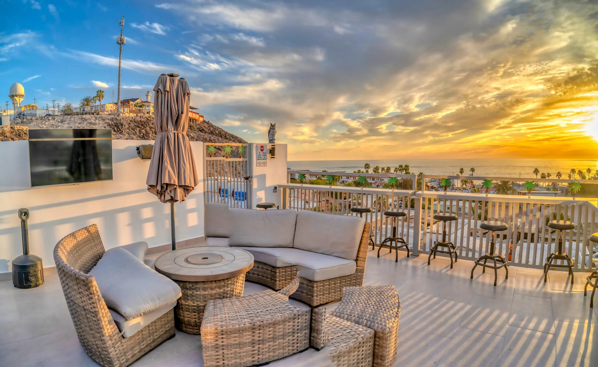 Terraza con muebles de ratán y cojines, vista al mar al atardecer, con palmeras y edificios en el fondo.
