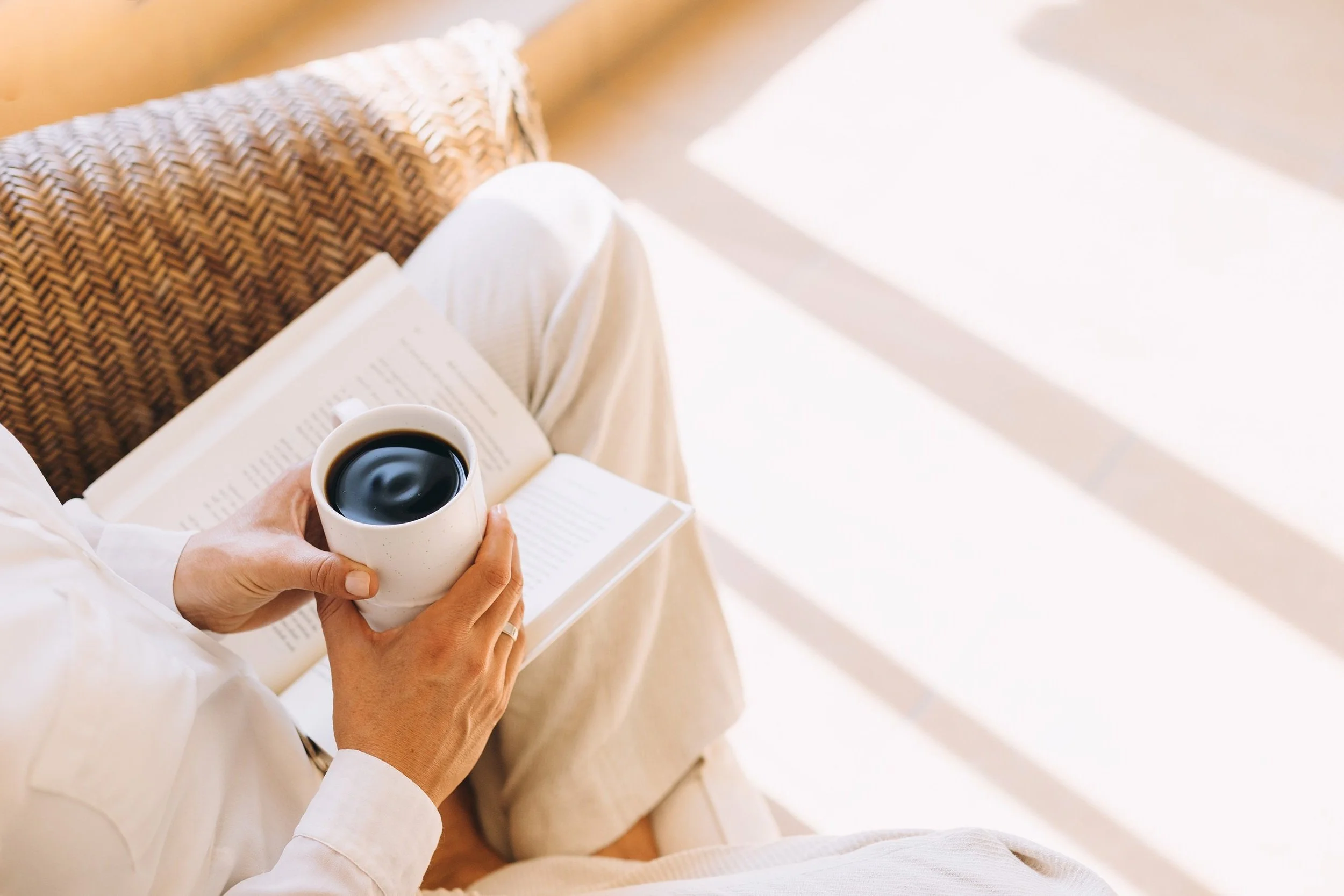 Person sitting in a chair holding a white mug filled with black coffee, with an open book on their lap, in a bright room with sunlight.