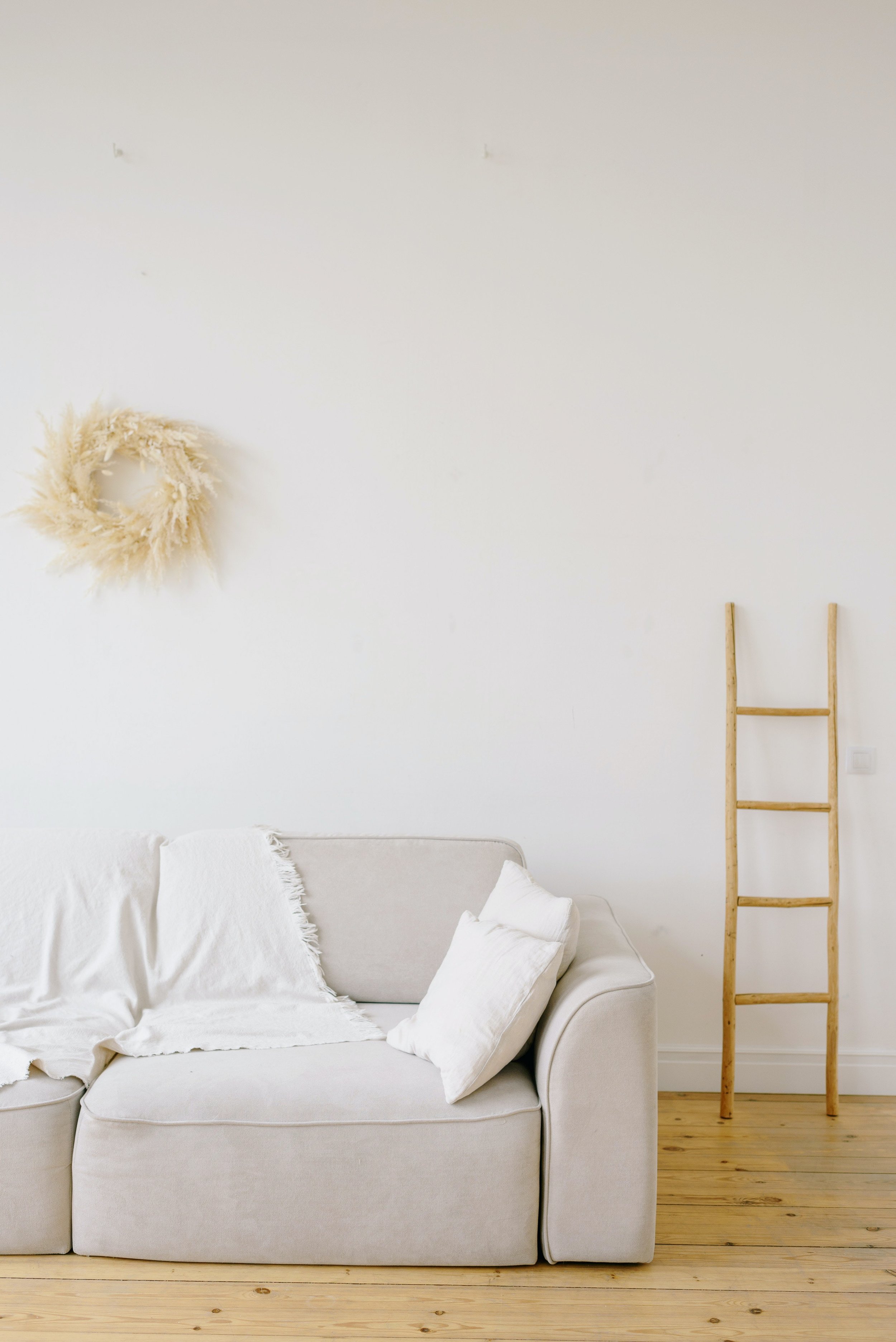 A white sofa with white pillows in a minimalist living room with wooden flooring. A ladder leaning against a white wall with a cream-colored fluffy wreath hanging on it.
