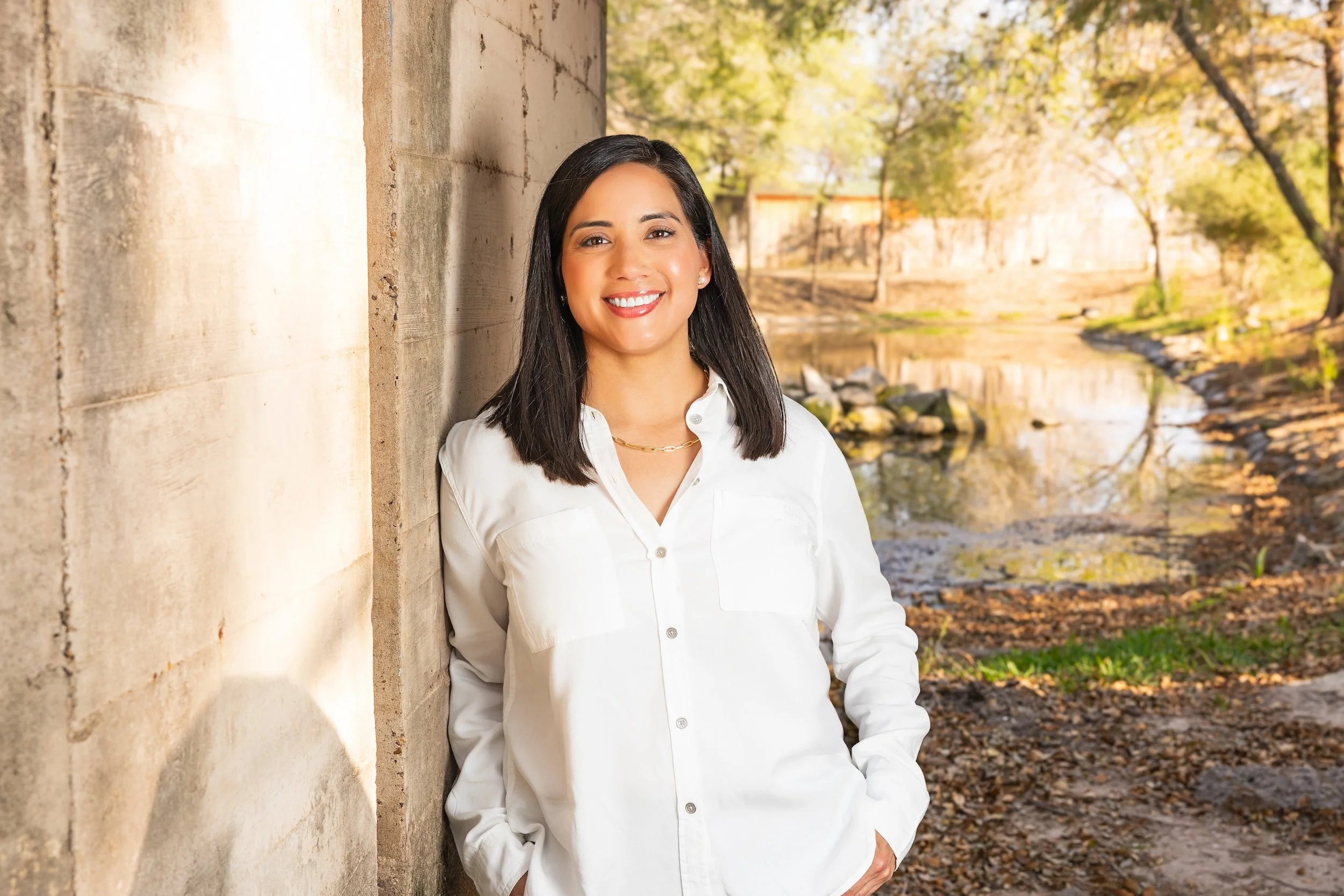 A woman with straight black hair, wearing a white button-up shirt and jewelry, leaning against a concrete wall outdoors near a river with trees in the background during fall.