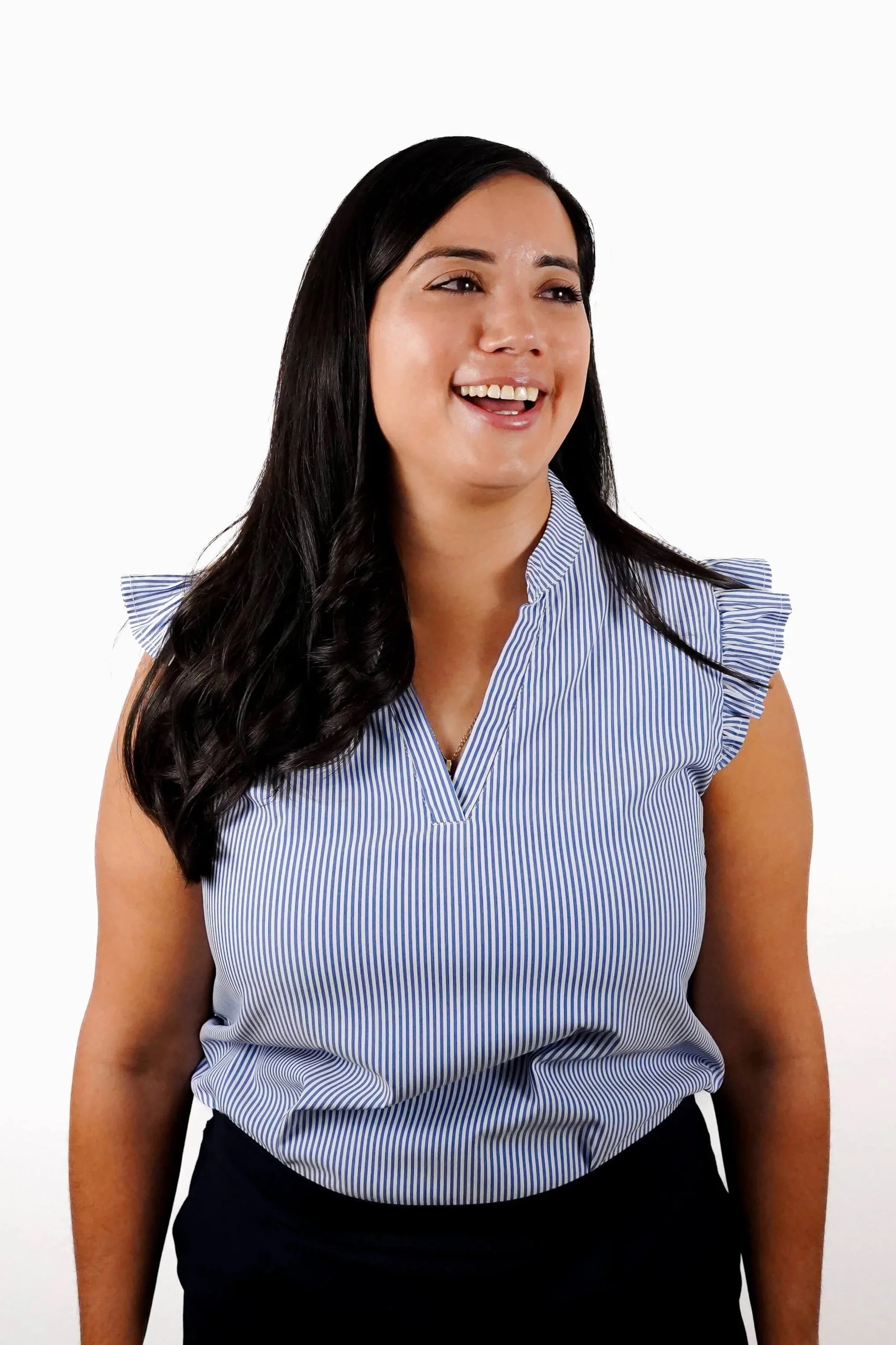 A woman with long dark hair smiling, wearing a sleeveless blue and white striped blouse and black pants, posed against a plain white background.