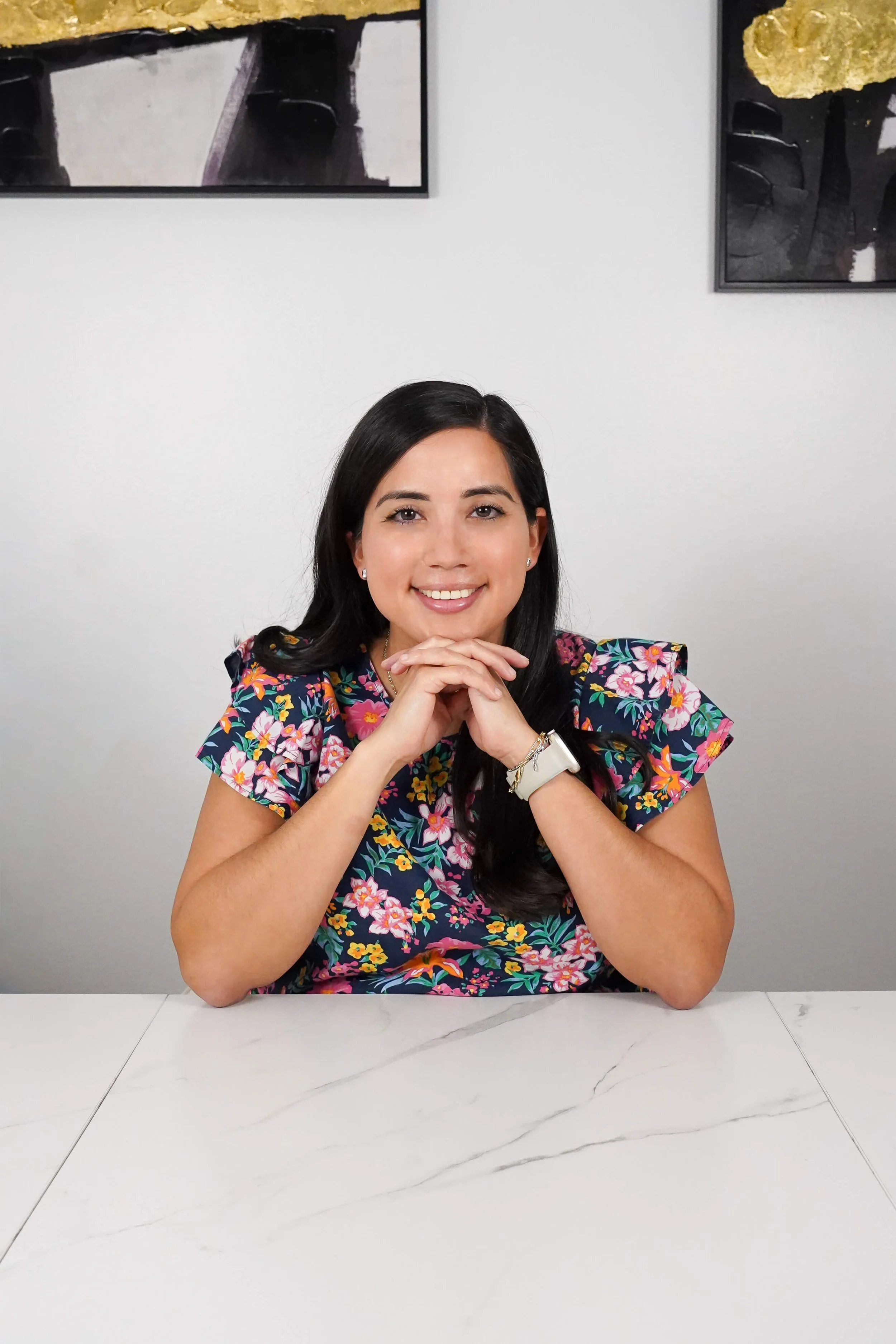 A woman with dark hair smiling at the camera, resting her chin on her hands, sitting at a marble table, wearing a colorful floral top, with framed artwork on the white wall behind her.