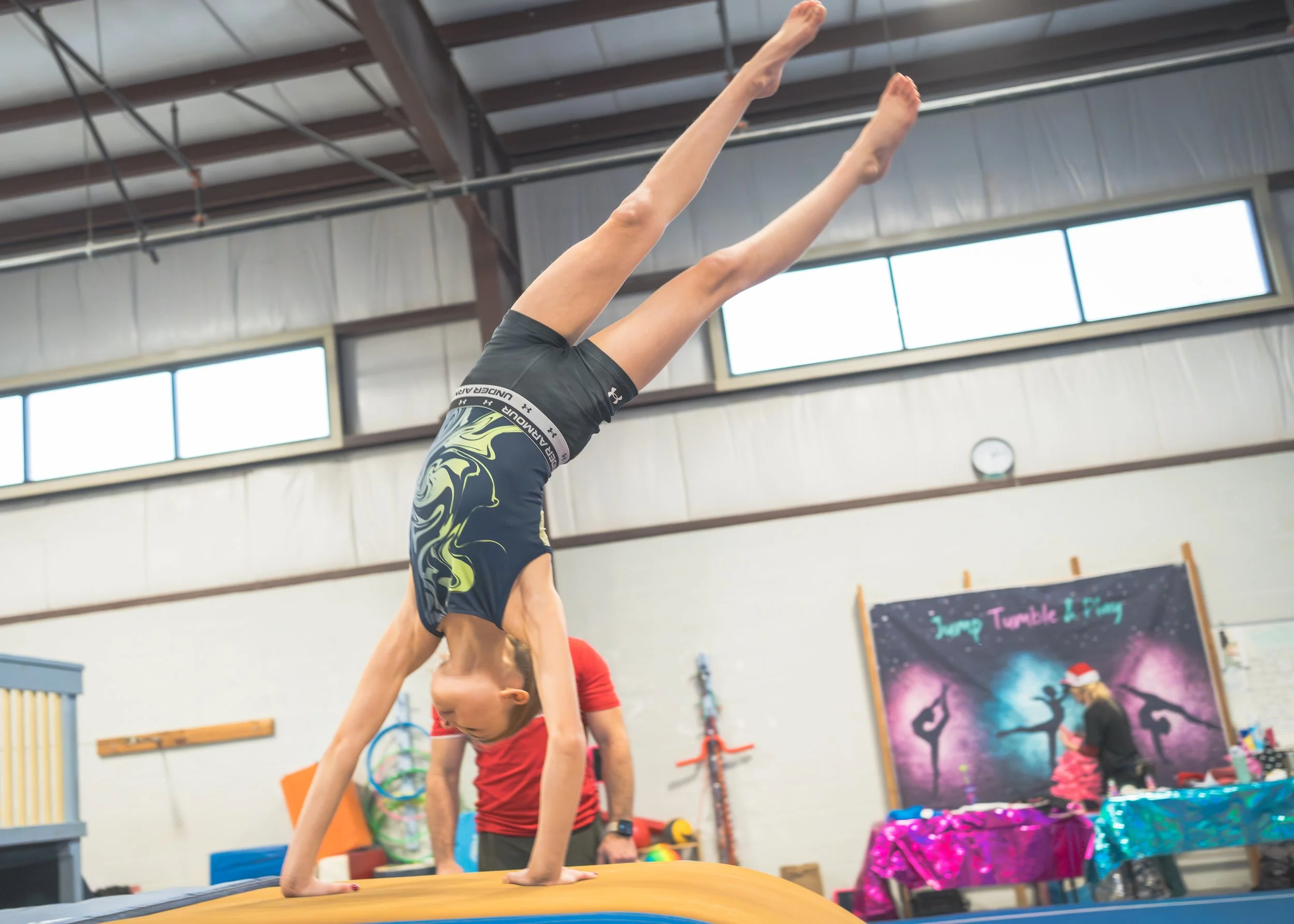 A girl performing a handstand on a tumbling mat in a gymnastics gym, with another person in the background and a colorful banner that reads 'Jump Tumble & Fly'.