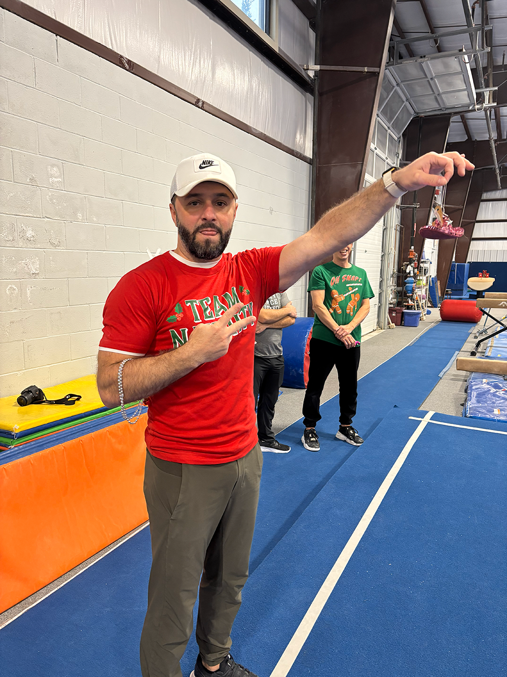 A man in a red Christmas-themed shirt and white Nike cap is making a peace sign with one hand and holding a small ornament or toy in the other in a gym or gymnastics training facility. He is standing on a blue mat, with other people and gym equipment visible in the background.