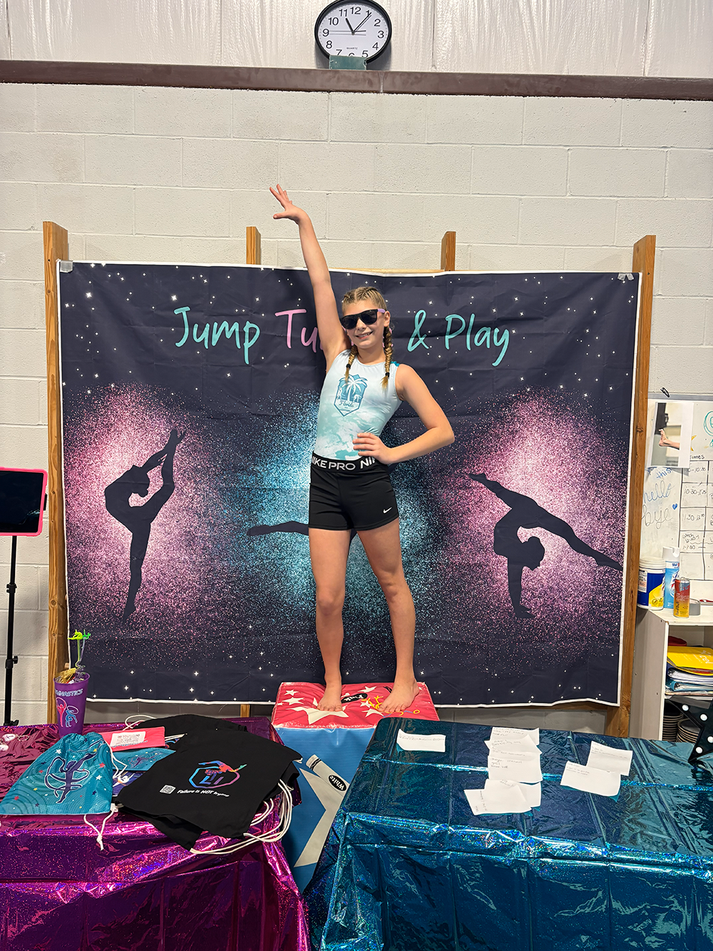 A girl in sunglasses and athletic wear poses on a pink and blue box in front of a birthday backdrop with silhouettes of gymnasts and the text 'Jump Turn & Play.' She is standing on the box with one arm raised above her head and a hand on her hip, smiling. The table in front has pink, blue, and black items, including a water bottle and T-shirts.