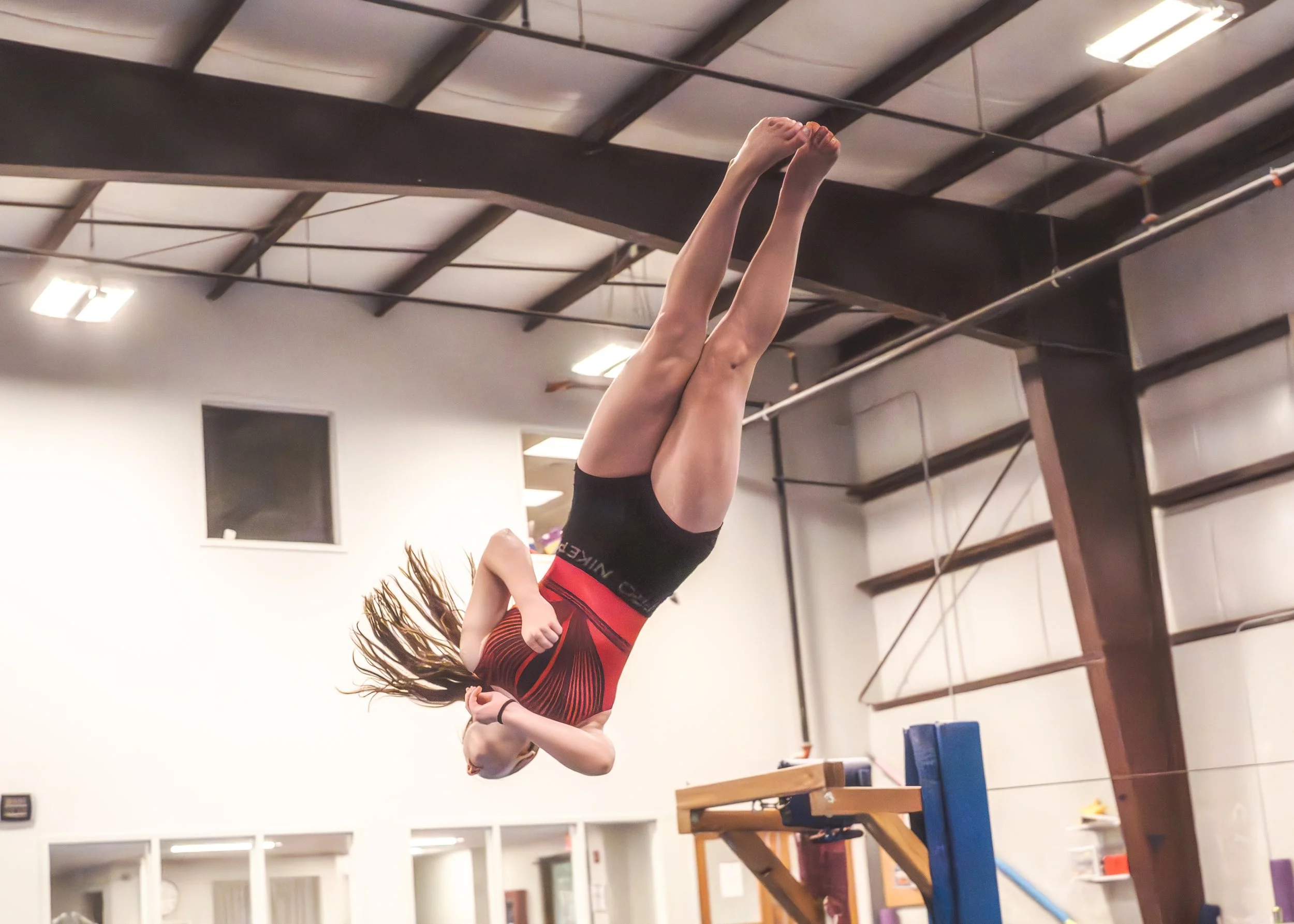 A female gymnast in a red and black leotard performing a flip on the balance beam in an indoor gymnastics facility.