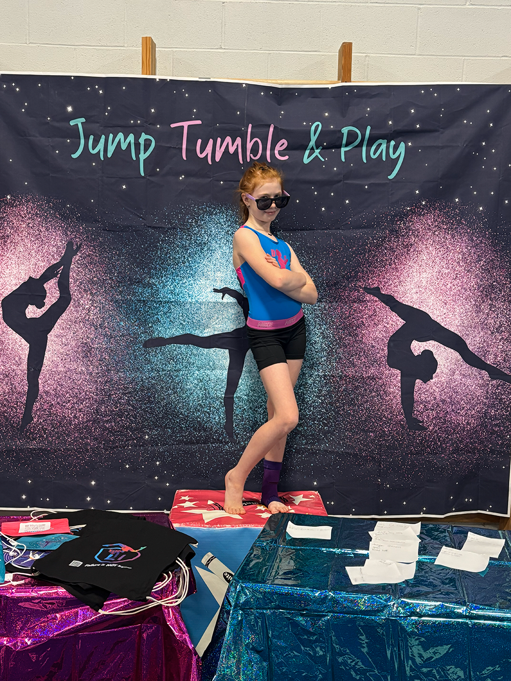 A young girl standing on a star-shaped platform with her arms crossed, posing in front of a backdrop that says 'Jump Tumble & Play' and features silhouettes of gymnasts in various poses, wearing sunglasses and a blue athletic outfit with a purple waistband.