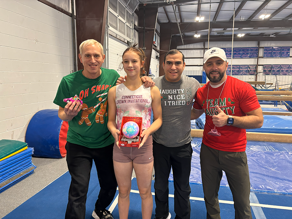 Four people standing inside a gymnasium, celebrating. A young girl in the center holding a glowing globe award, flanked by three men. The man on the left wearing a green holiday shirt, the man next to her wearing a gray t-shirt, and the man on the right wearing a red shirt and white cap, holding a glass. The gym has balance beams and fitness equipment in the background.