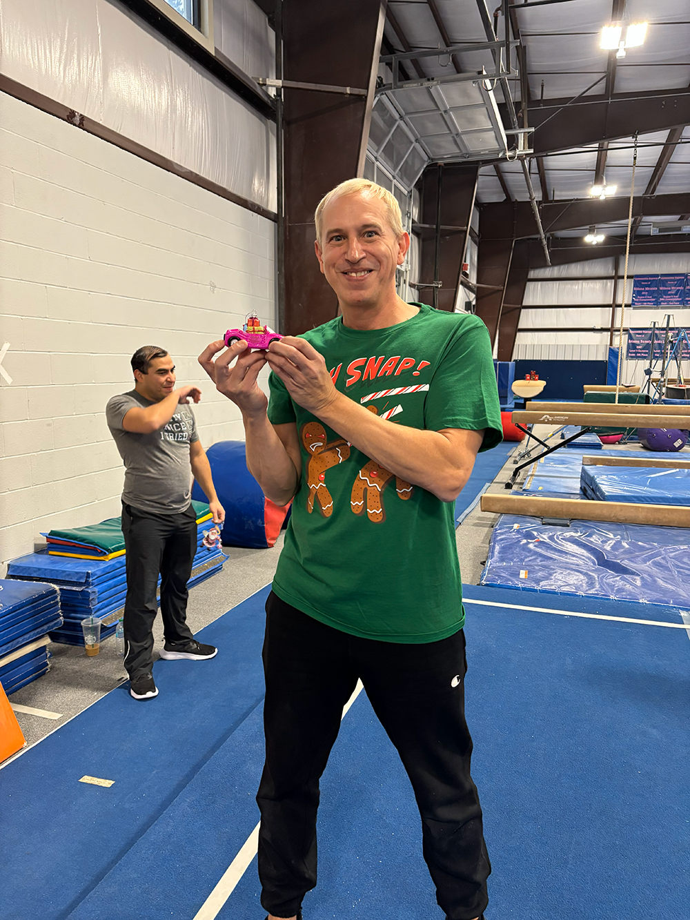 A man with short gray hair wearing a green Christmas-themed shirt and black pants is smiling and holding a small pink toy car with a decorated gingerbread man on top. He is in an indoor gym with blue mats, gymnastics equipment, and another man in the background.