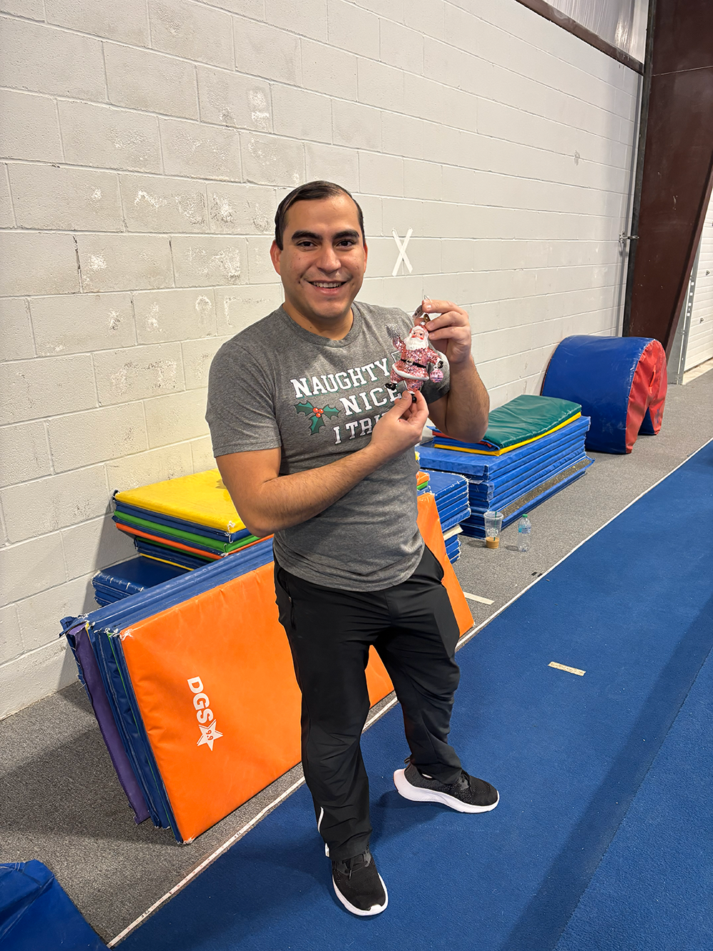 A man standing in a gymnasium holding a Christmas ornament of Santa Claus, smiling at the camera, with colorful mats and foam padding stacked against the wall behind him.