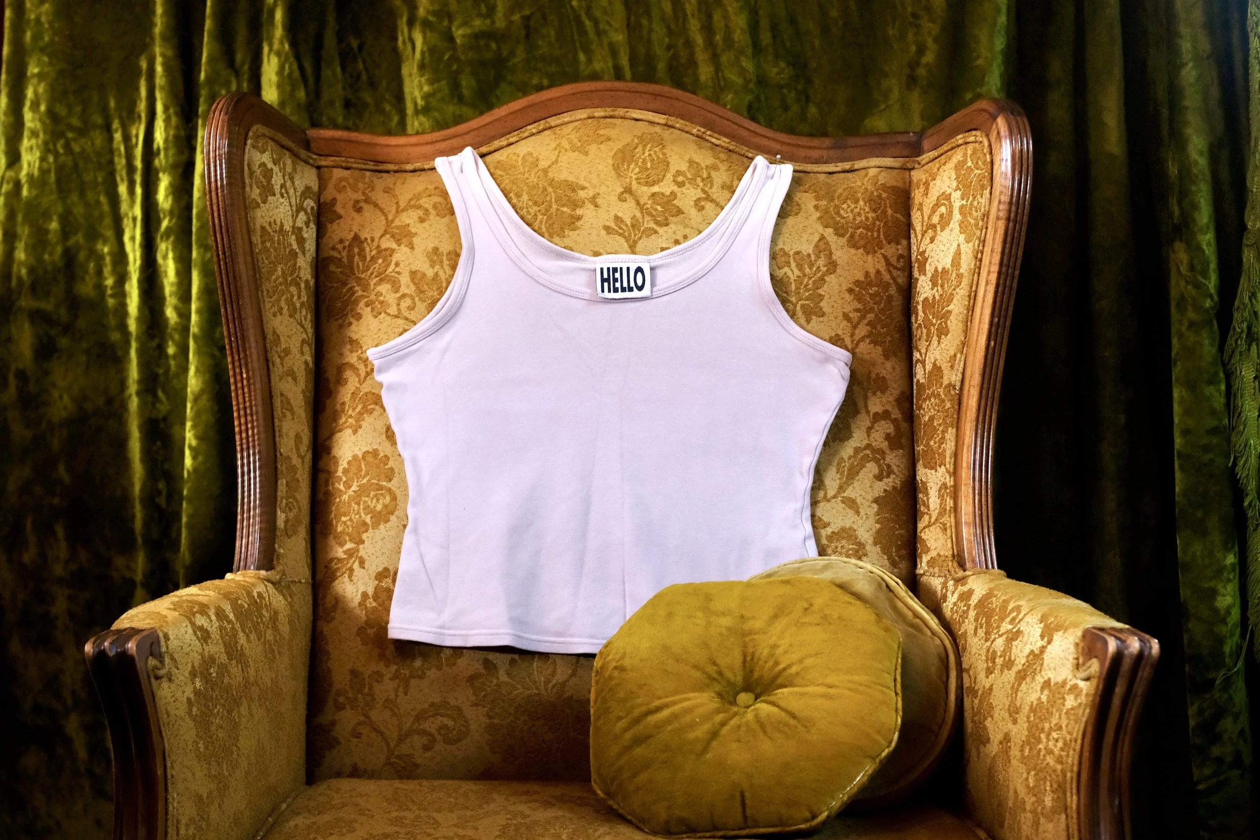 A white sleeveless tank top with a 'HELLO' label displayed on a vintage yellow and gold patterned armchair, with a round mustard-colored cushion in front of it, in front of green curtains.