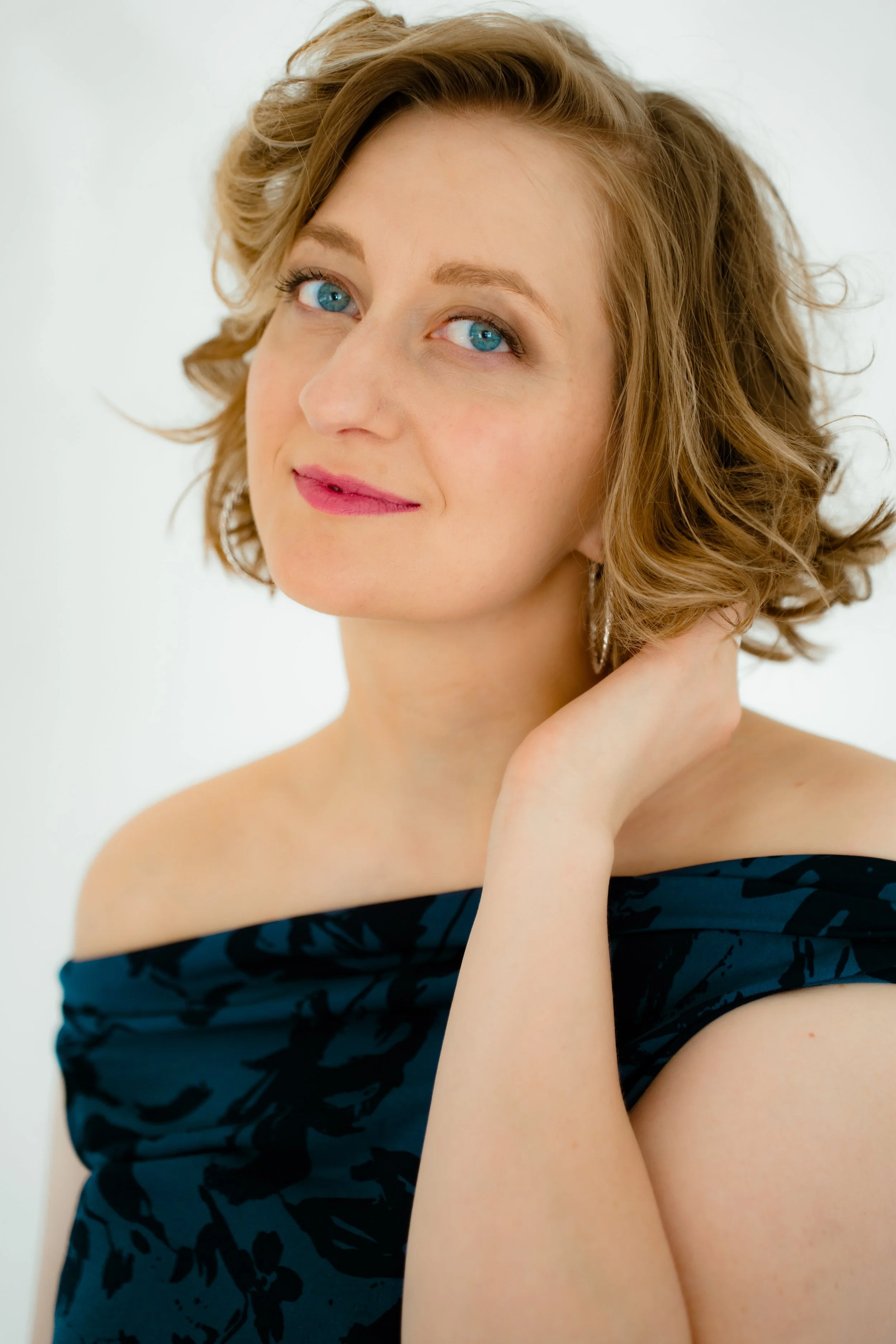 A woman with short wavy blonde hair, blue eyes, wearing a dark off-shoulder top with a floral pattern, resting her left hand on her neck, smiling softly against a plain light background.