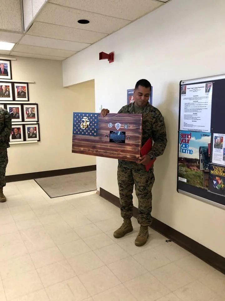 A military service member in camouflage uniform holding a large framed American flag with military insignia while standing in a hallway near a bulletin board, with another person partially visible on the left side of the image.