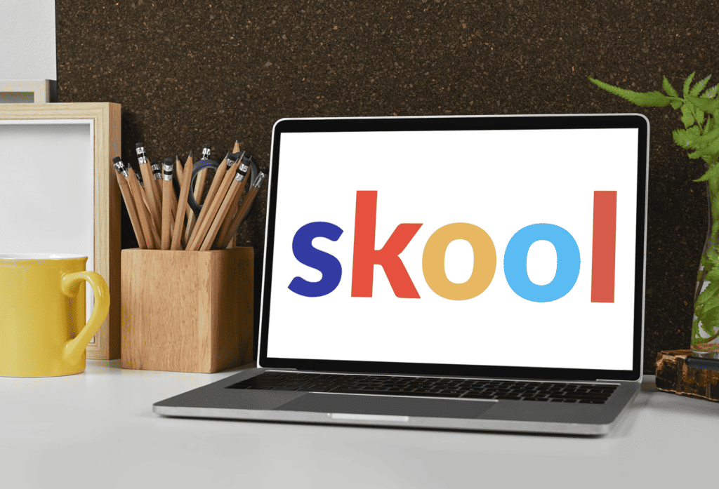 Laptop on desk displays the word 'skool' in colorful letters, next to a yellow mug, a wooden container with pencils and scissors, and a potted plant against a cork board background.