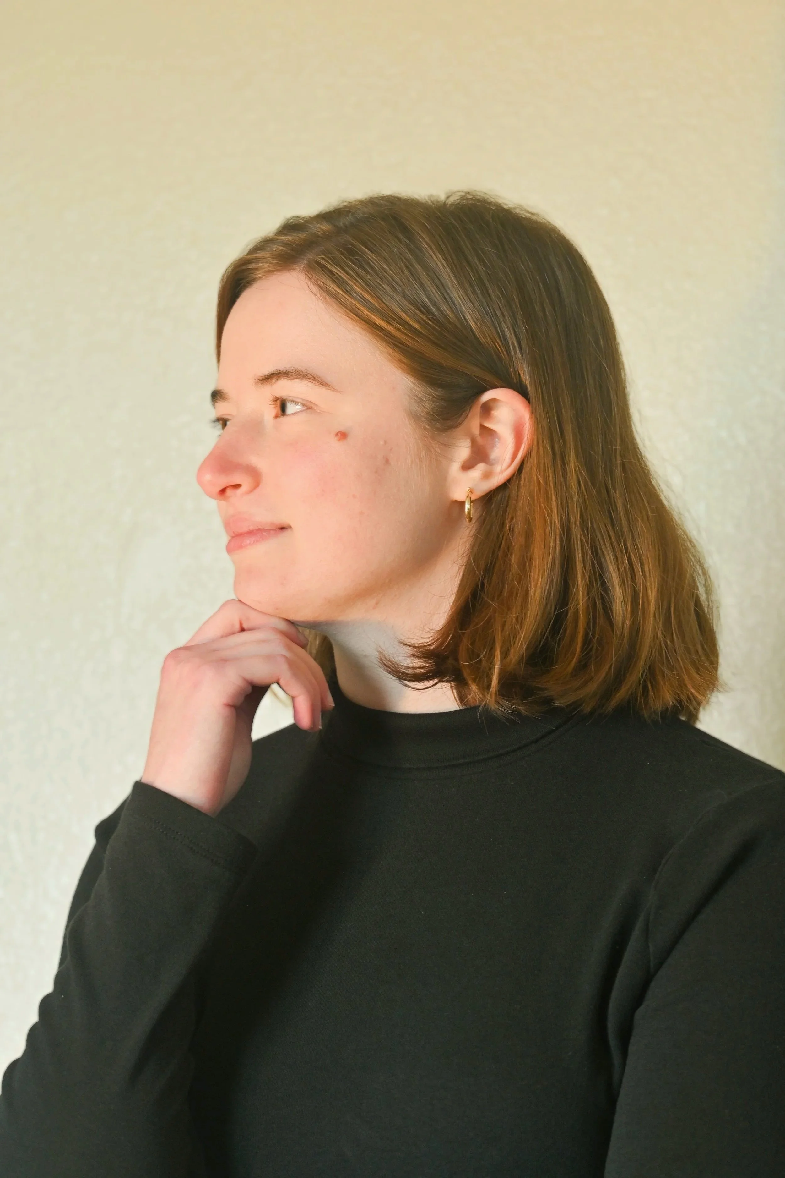 A young woman with shoulder-length brown hair, wearing a black turtleneck, is resting her chin on her hand and looking to her left, in front of a beige textured background.