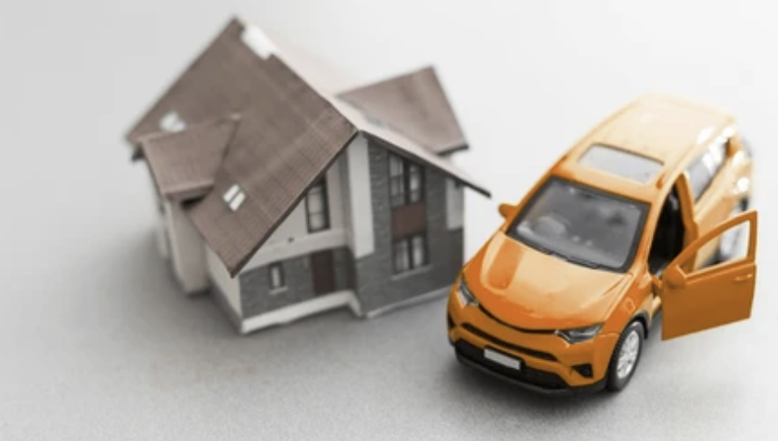 A miniature model house next to an orange toy car with an open door, placed on a plain surface.