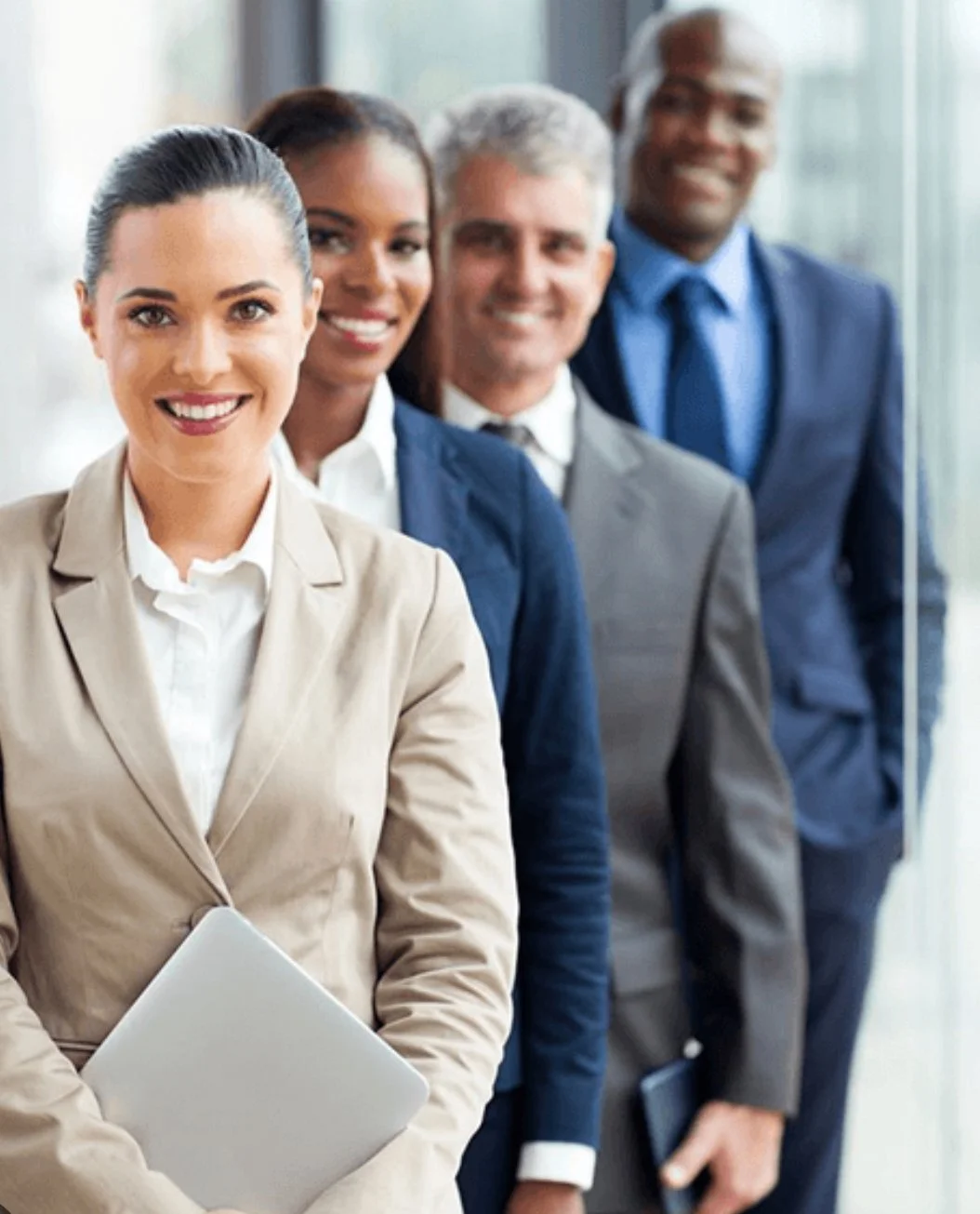 Four diverse professionals in business attire standing in a line, smiling in an office setting.