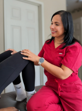 A healthcare worker in red scrubs smiling and working with a patient, holding the patient's leg during a medical treatment or examination.