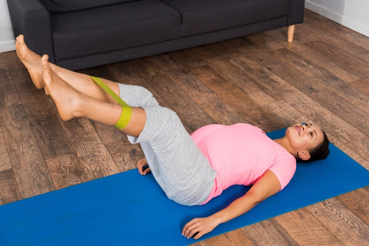A woman lying on her back on a blue yoga mat with her legs raised and secured with resistance bands, smiling during an exercise in a living room with wooden floors and a dark gray couch.