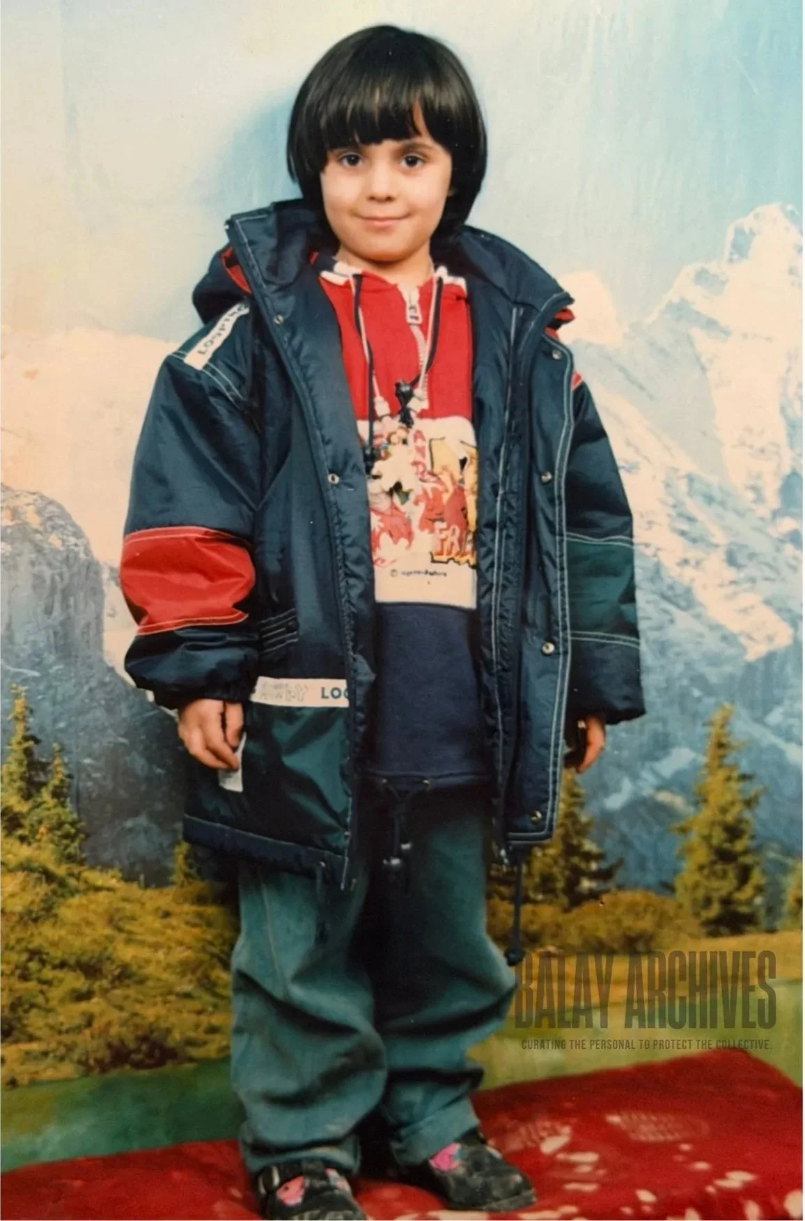 A young boy with dark hair wearing a blue and red winter jacket, a red hoodie with a graphic design, gray pants, and sneakers, standing on a red carpet with a mountain landscape background.