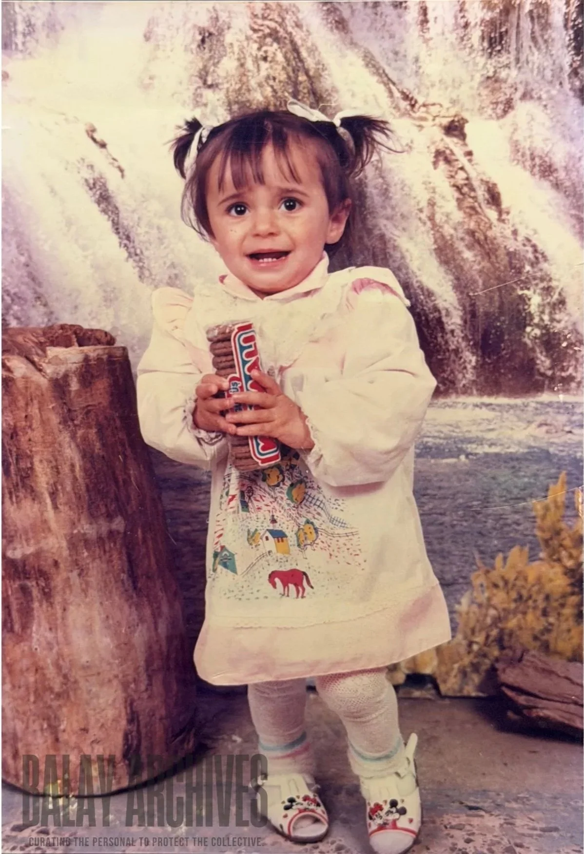 Young girl with pigtails holding a stack of cookies, standing in front of a waterfall background, wearing a white dress with colorful embroidery, pink tights, and Mickey Mouse slippers.