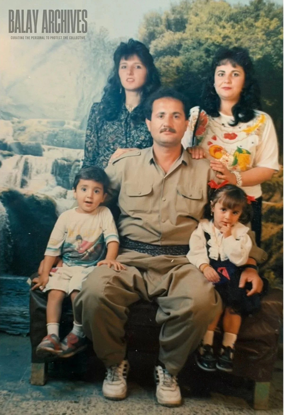 A family portrait of a man with four children in front of a scenic backdrop of a waterfall and trees. The man is sitting with two children on his lap and two women standing behind him.