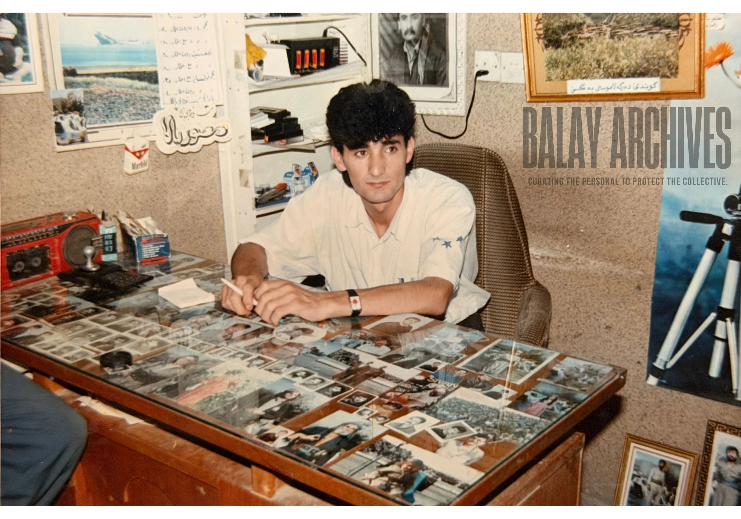 A man sitting at a cluttered desk covered with photographs in an office. There are posters and pictures on the wall behind him, including a portrait of a man with a mustache, a landscape photo, and a butterfly. The man is holding a pen, wearing a white short-sleeved shirt with a star emblem on the sleeve, and has black curly hair.