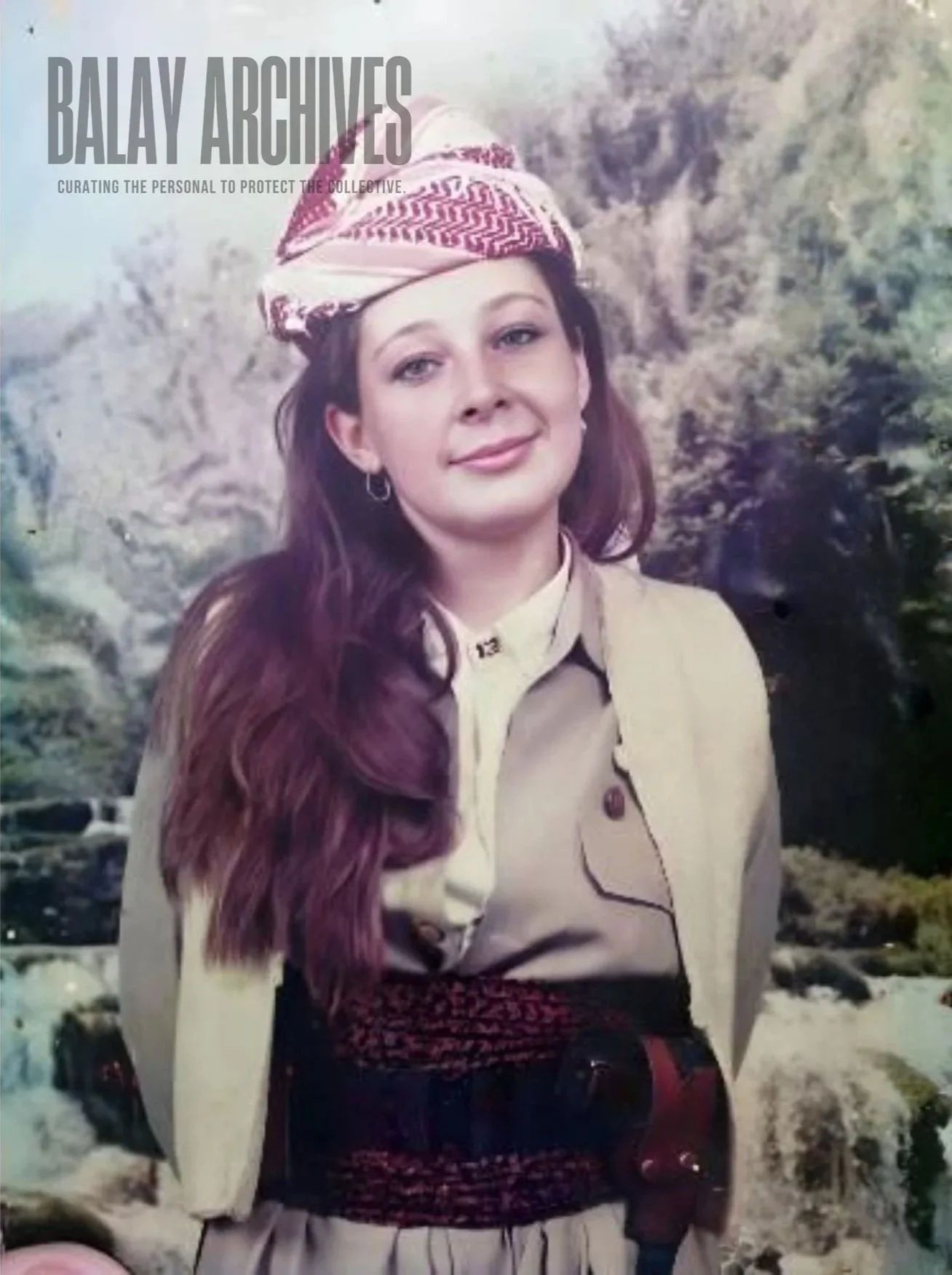A woman with long, wavy, auburn hair wearing a beige uniform with a necktie, earrings, and a headscarf with a red and white pattern, standing outdoors with a rocky background.