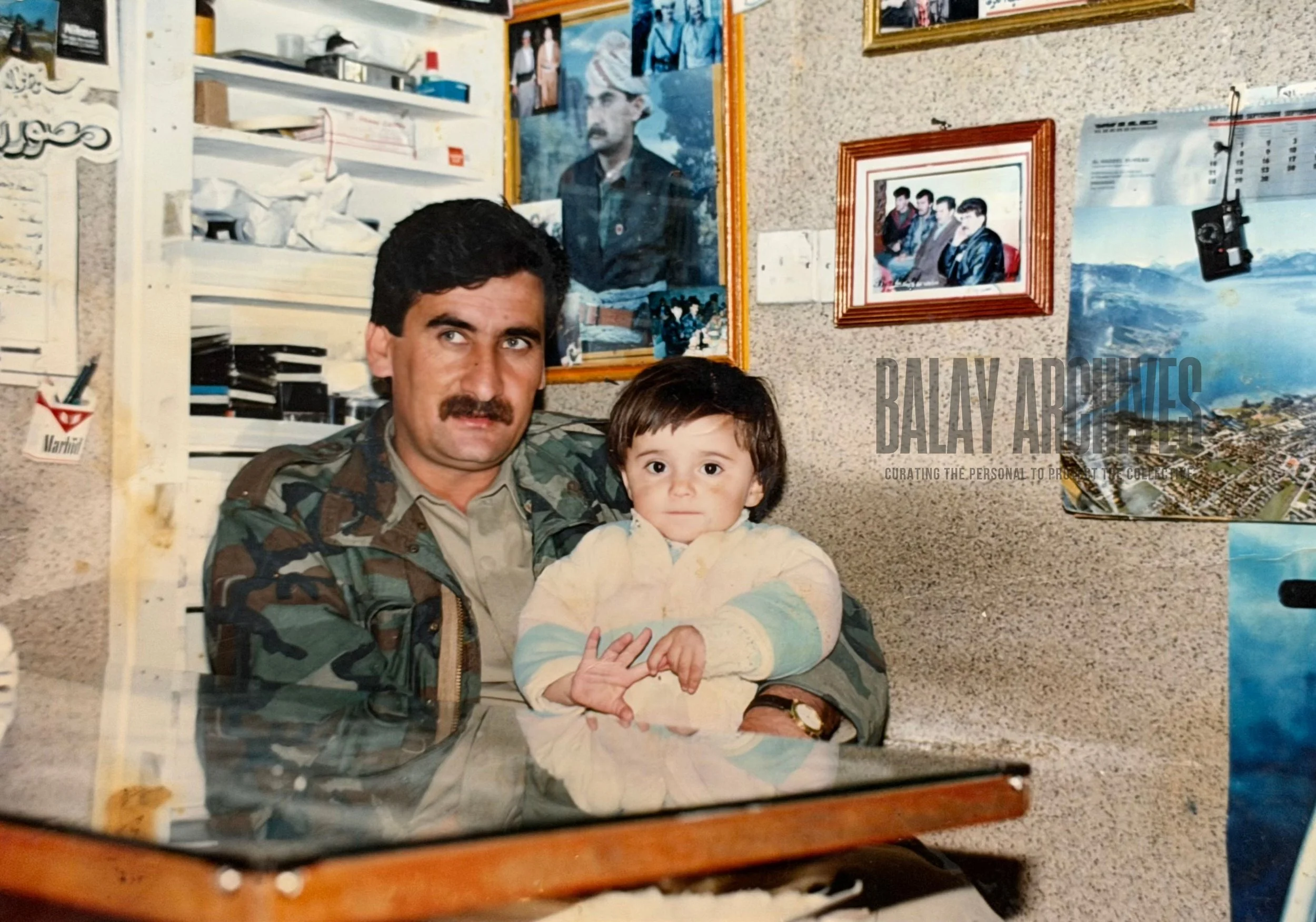 A man in a military uniform holding a young child in an indoor setting decorated with photographs and posters on the wall.