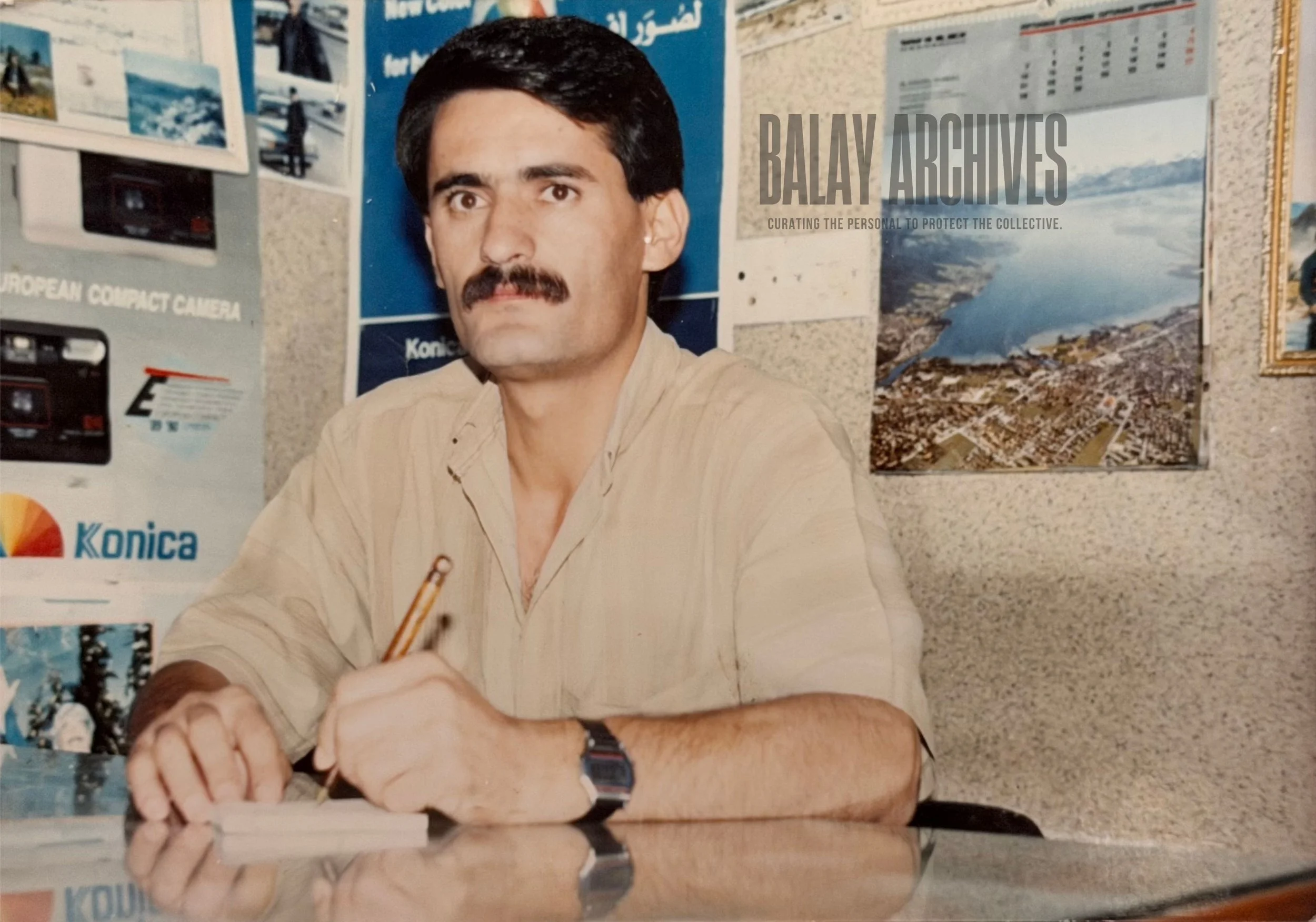 A man with dark hair, a mustache, wearing a beige shirt and a wristwatch, sitting at a desk with a pen in his hand. Behind him are posters and photographs on a bulletin board, including images of cameras and a large poster of a landscape with water and mountains.