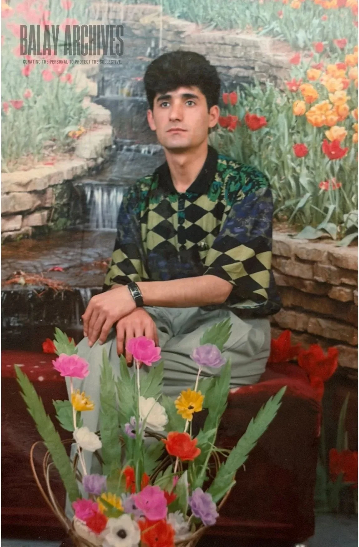 A young man with dark hair and a serious expression sitting on a bench with a floral bouquet in front of him. The background features a painted waterfall scene with flowers and greenery. The photo has a logo in the top left corner reading 'BALAY ARCHIVES' with a tagline below.