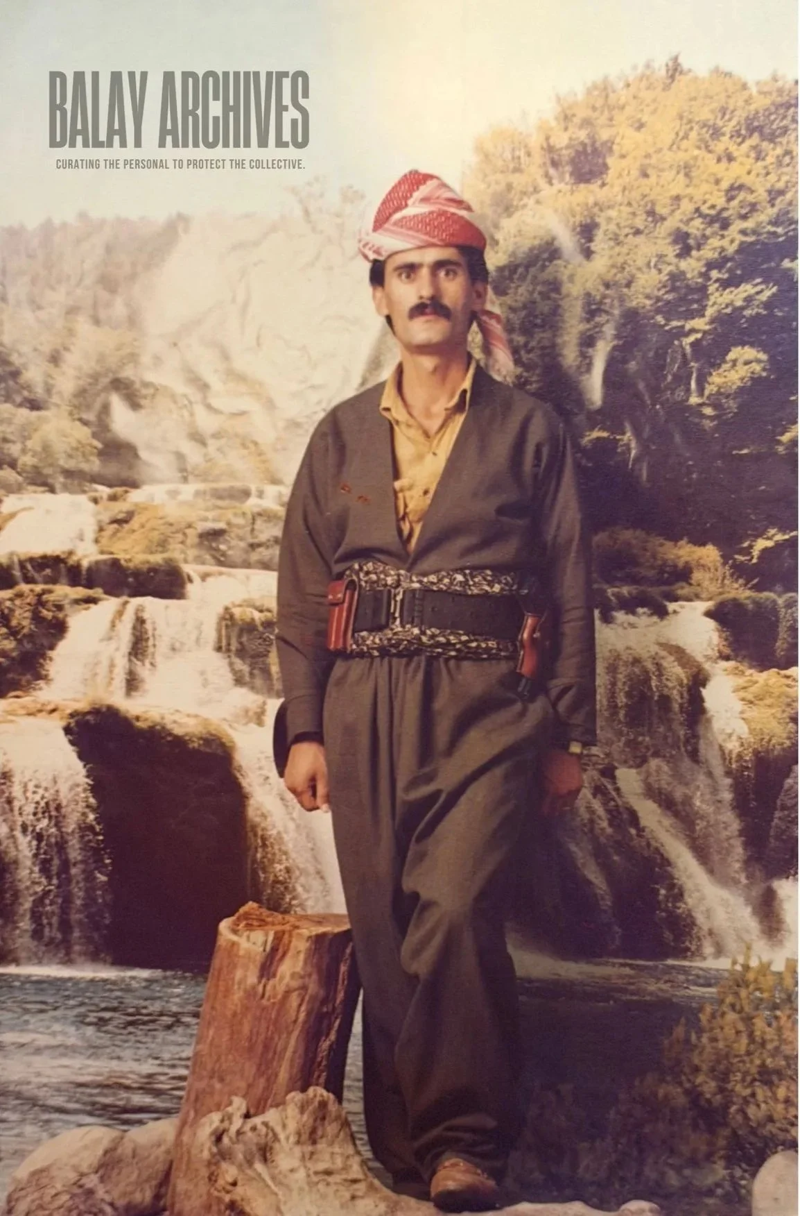 A man dressed in traditional Kurdish attire standing behind backdrop of waterfall and forested landscape in the background. He is wearing a headscarf, a dark robe, a yellow shirt, and a wide belt, with a serious expression.