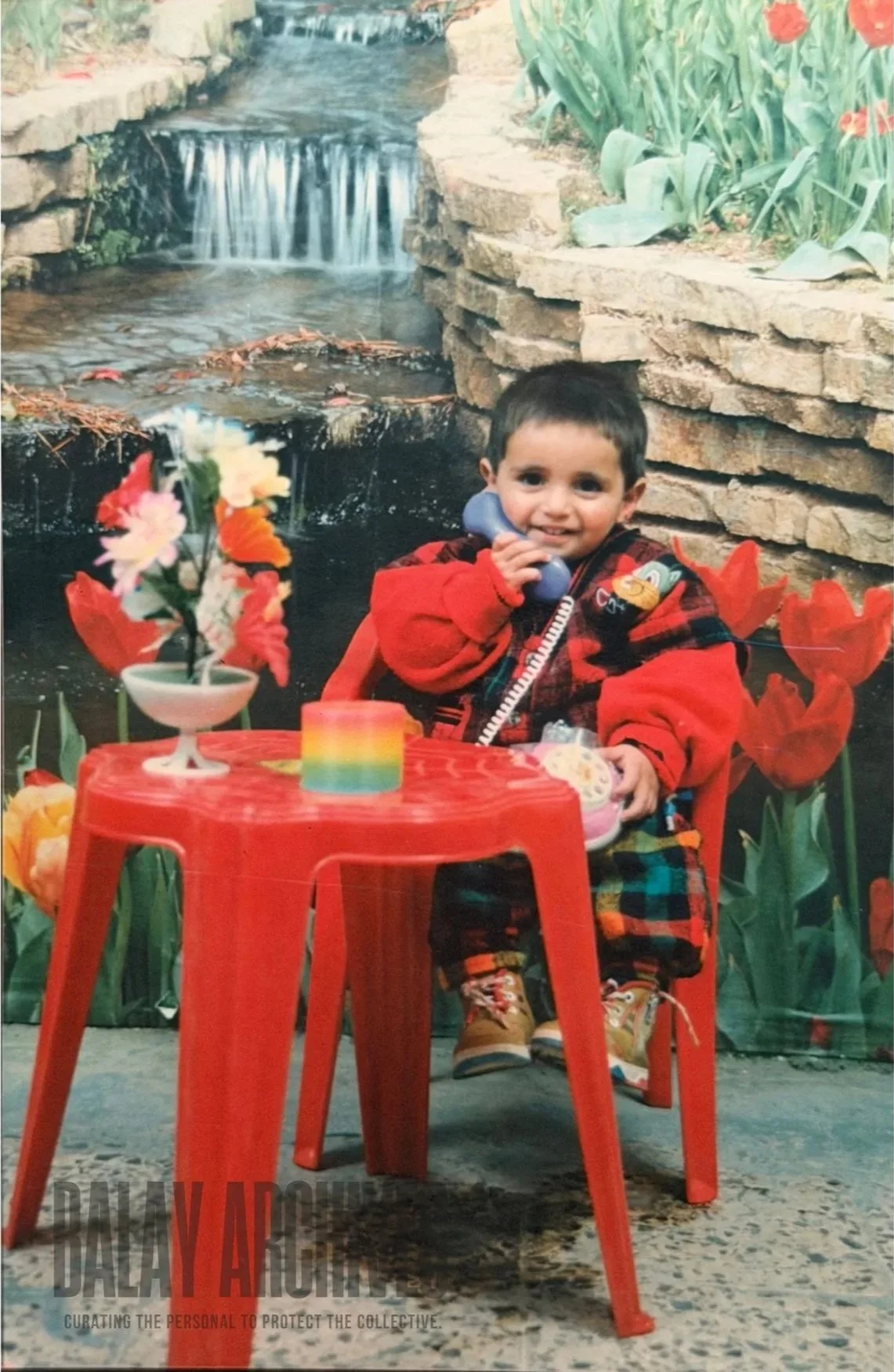 A young boy with dark hair and a red jacket sitting at a small red plastic table, holding a blue toy phone to his ear with a smile. The table has a colorful rainbow paper or fabric at the center and a small white vase with pink and white flowers. Behind him is a backdrop with a waterfall and stone wall, surrounded by large red and yellow tulips and green foliage, mimicking a garden scene.