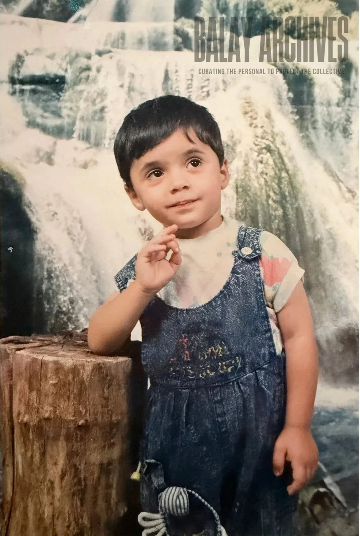 A young boy with black hair and brown eyes standing in front of a waterfall backdrop, resting his arm on a wooden log, and touching his chin thoughtfully.