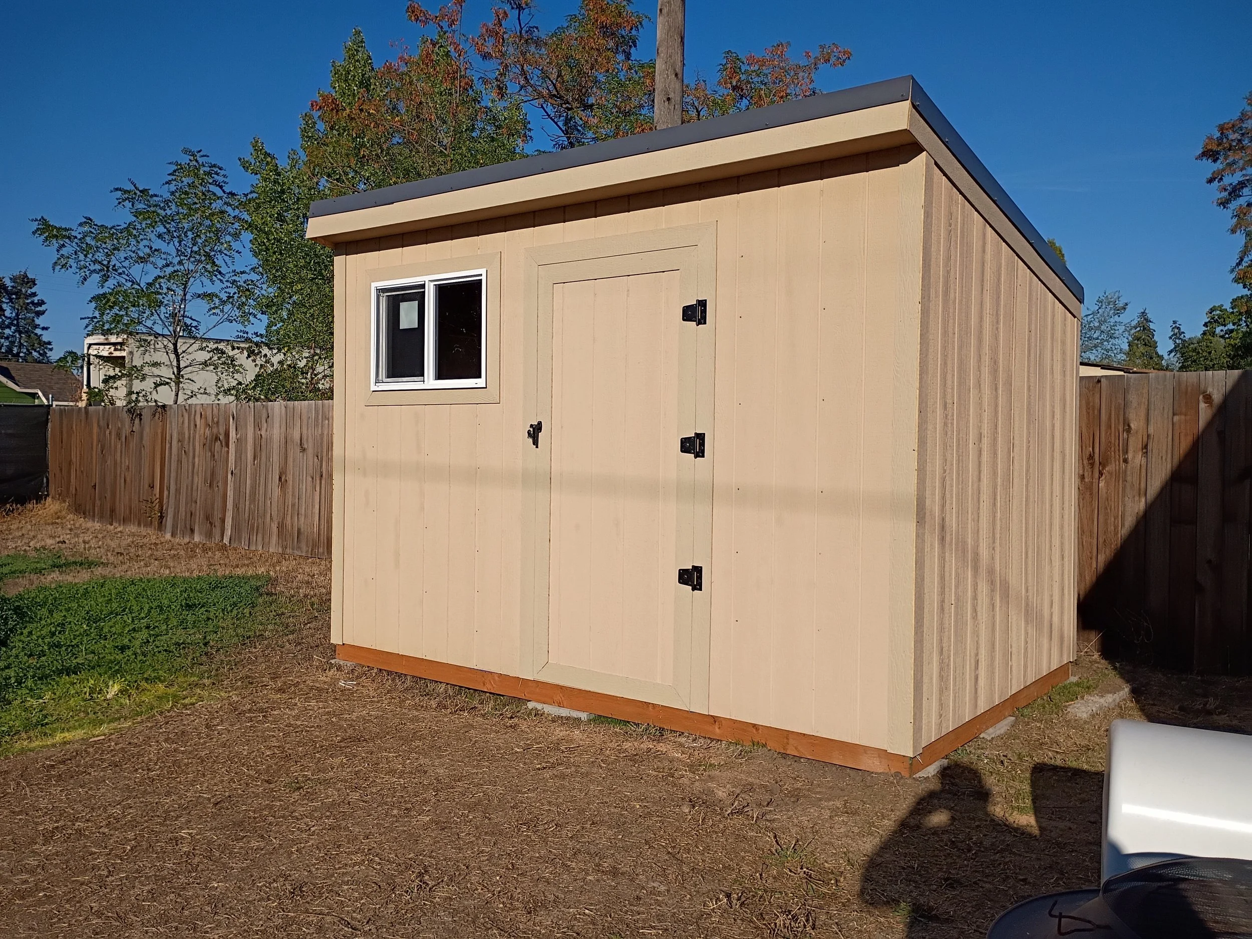A small, beige wooden shed with a sloped roof, a window with white trim, and a double door with black hinges and latch, situated in a backyard with a wooden fence and trees in the background.
