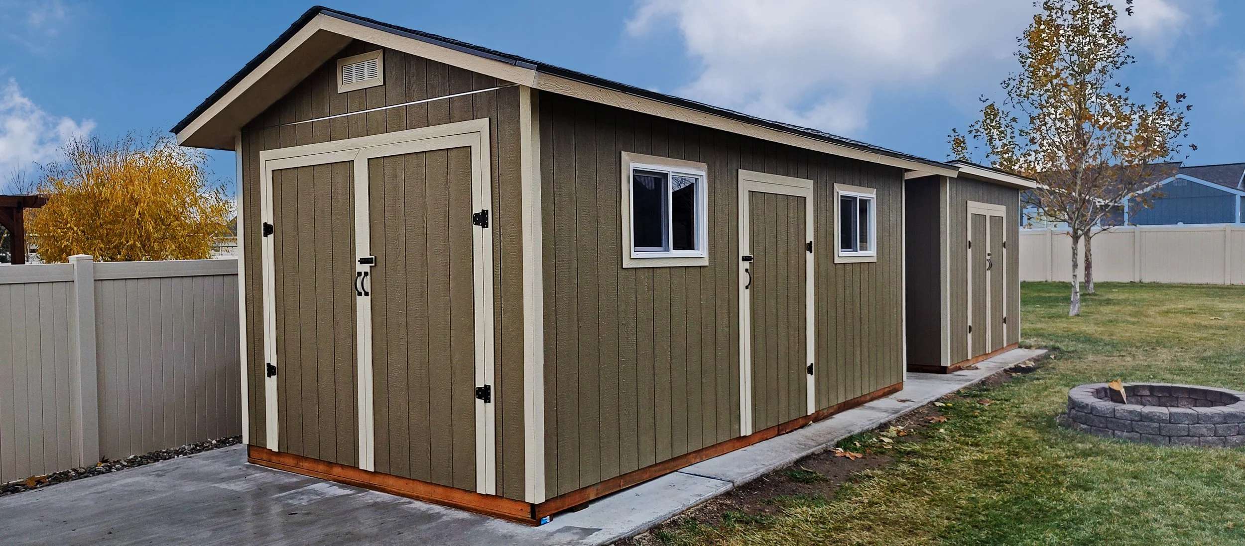 A small tan storage shed with white trim, three windows, and double doors in a backyard with a white fence and a tree with autumn leaves.