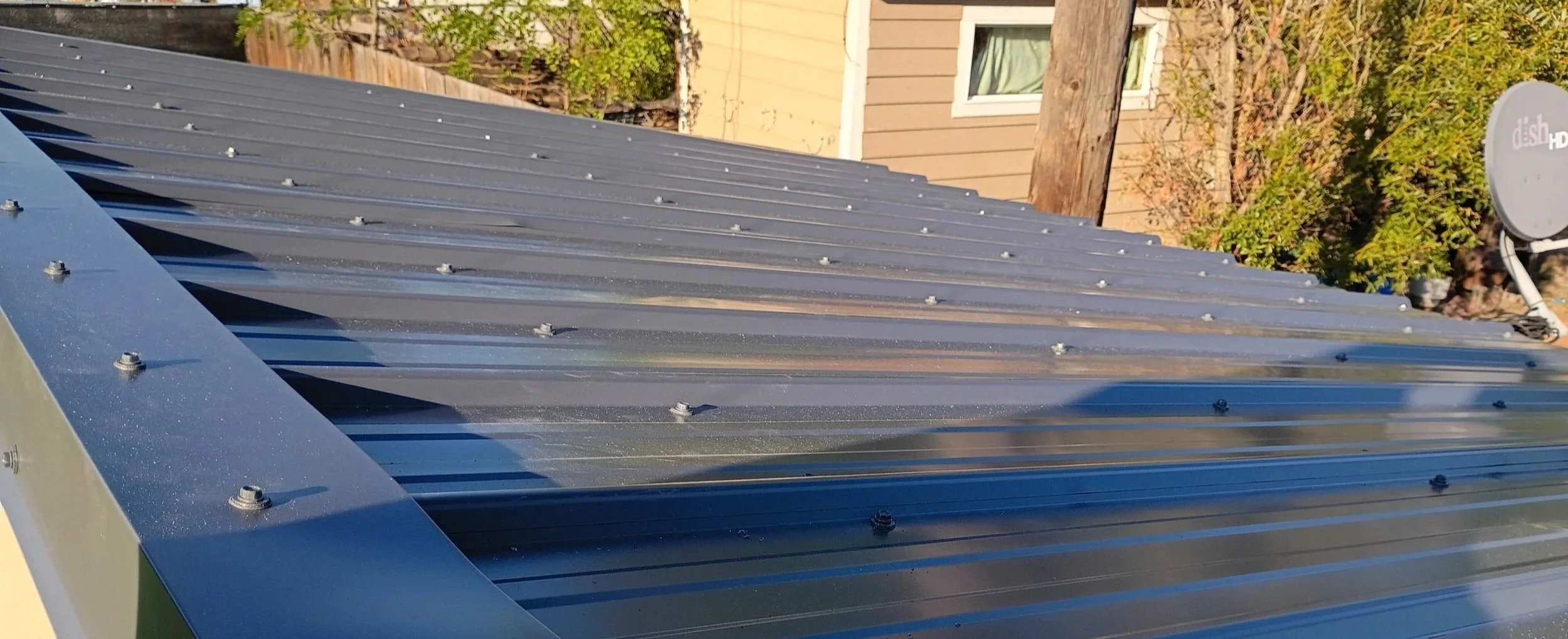 Close-up of a metal roof with visible screws, with a satellite dish and a house in the background.