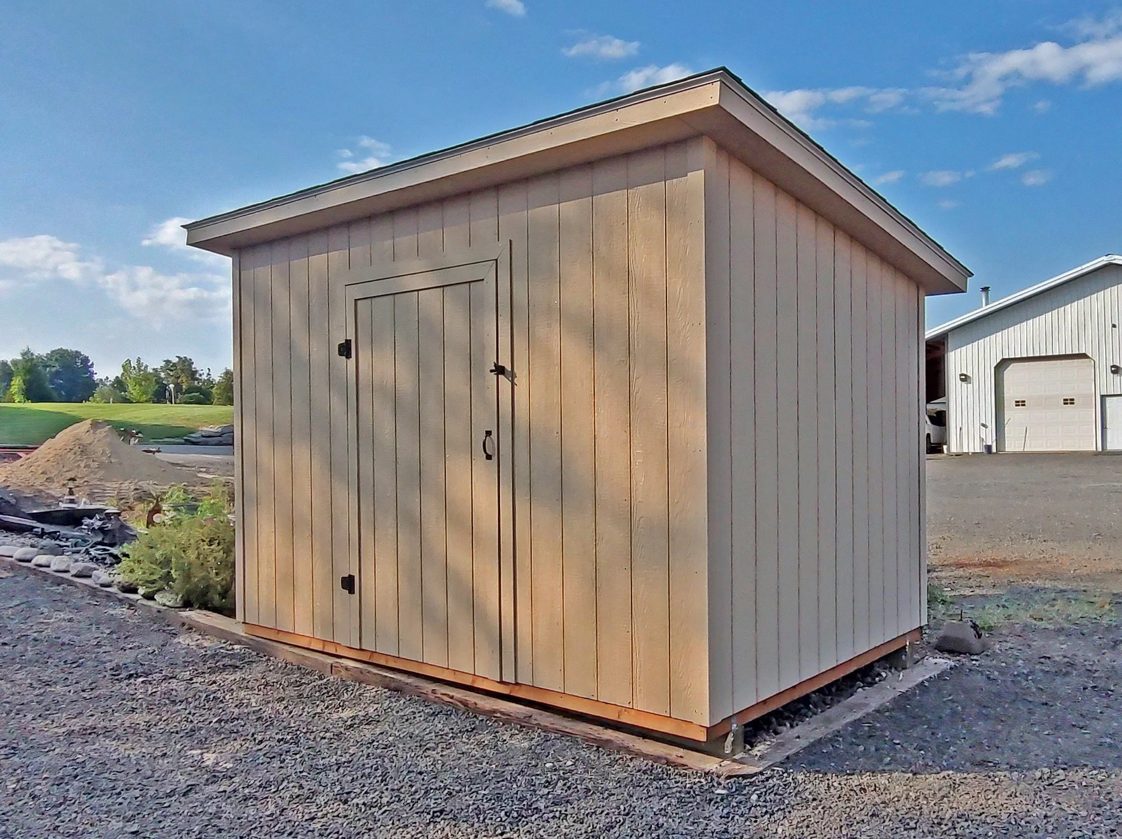 A small wooden shed with a slanted roof sits on a gravel area outdoors, with a white building and a grassy field in the background.