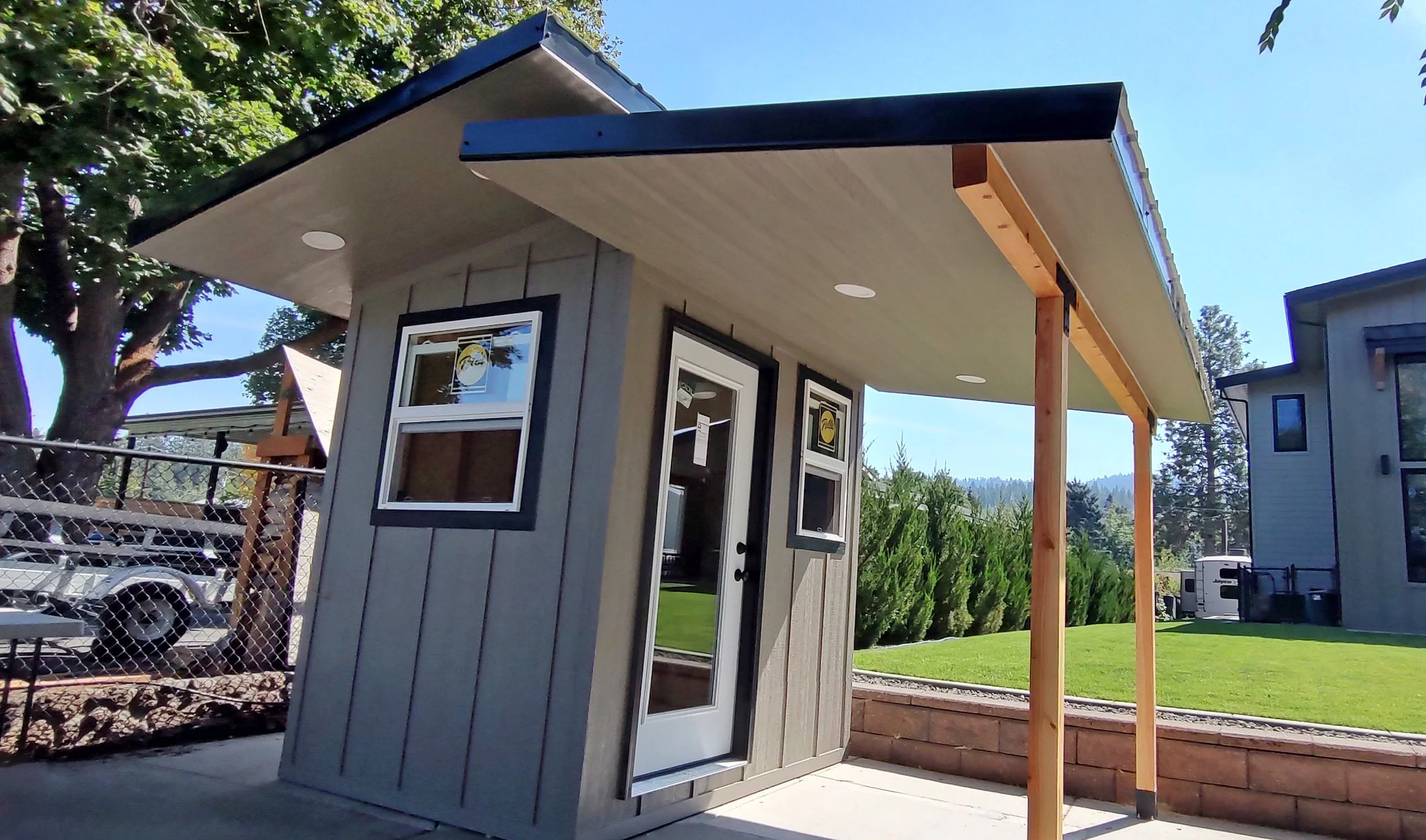 Small gray building with a sloped roof, two windows, and a door, under a larger roof extension with round ceiling lights; construction post supports the extension in a backyard with green grass and trees.