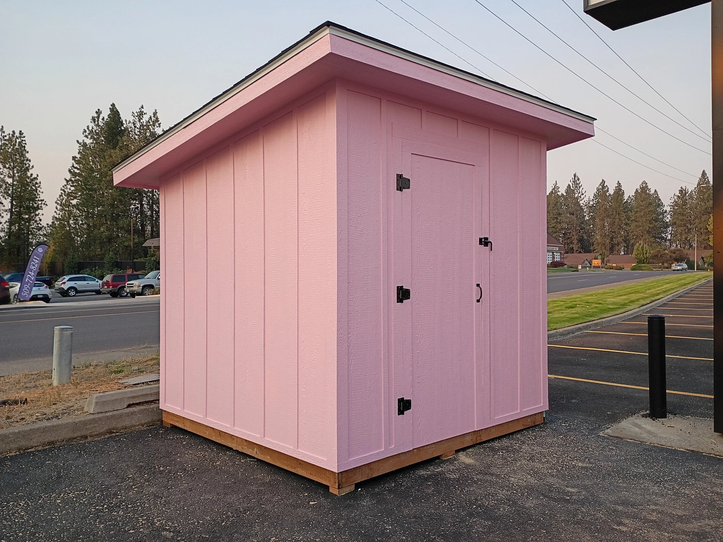 A small pink storage shed with black hinges and a handle, situated on a paved parking lot near a grassy area with trees in the background.