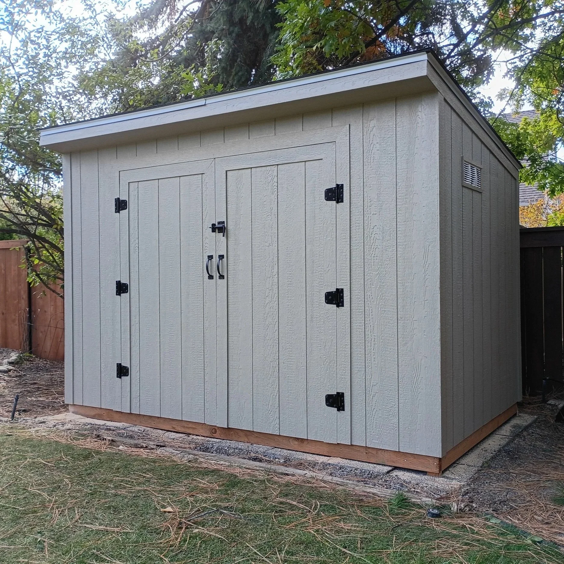 A small outdoor storage shed with beige vertical siding, black hinges, and handles, situated on a yard with grass and dirt. There is a tree with green foliage nearby and a black fence in the background.