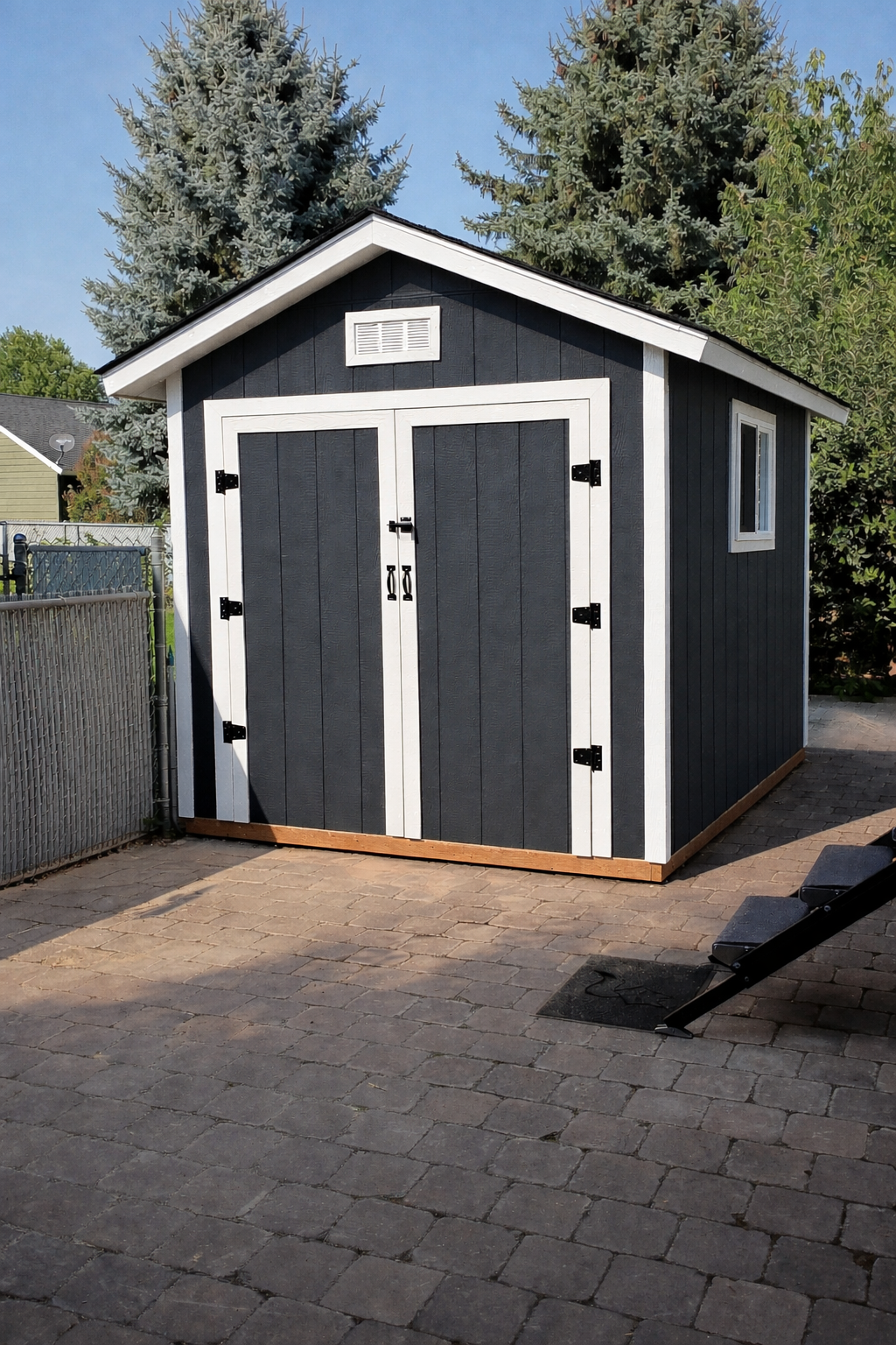A small black shed with white trim and double doors, located on a paved backyard patio, with a window on the right side and trees in the background.
