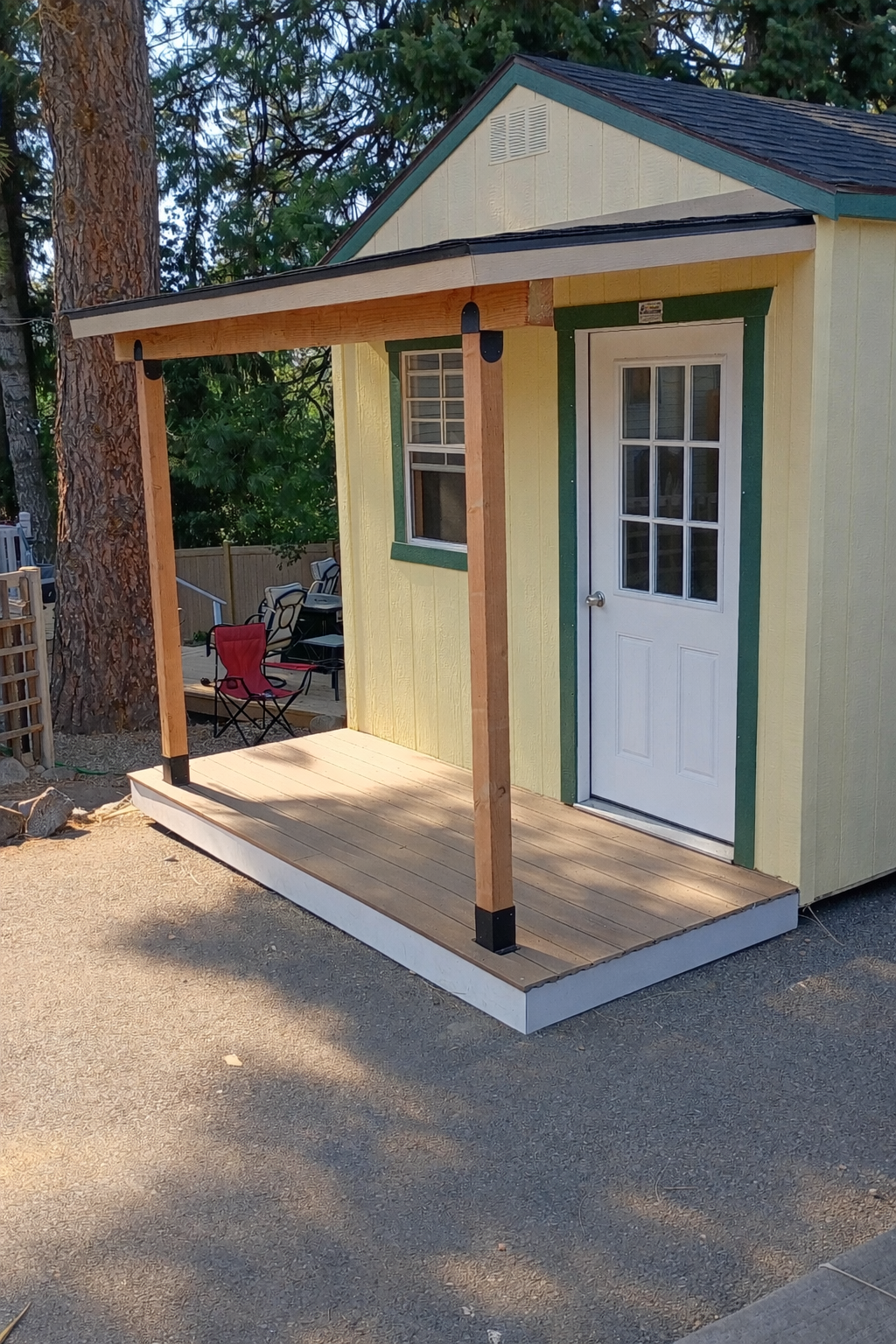 A small yellow shed with green trim and a white door with windowpanes, featuring a newly constructed wooden porch with a roof supported by four wooden posts.