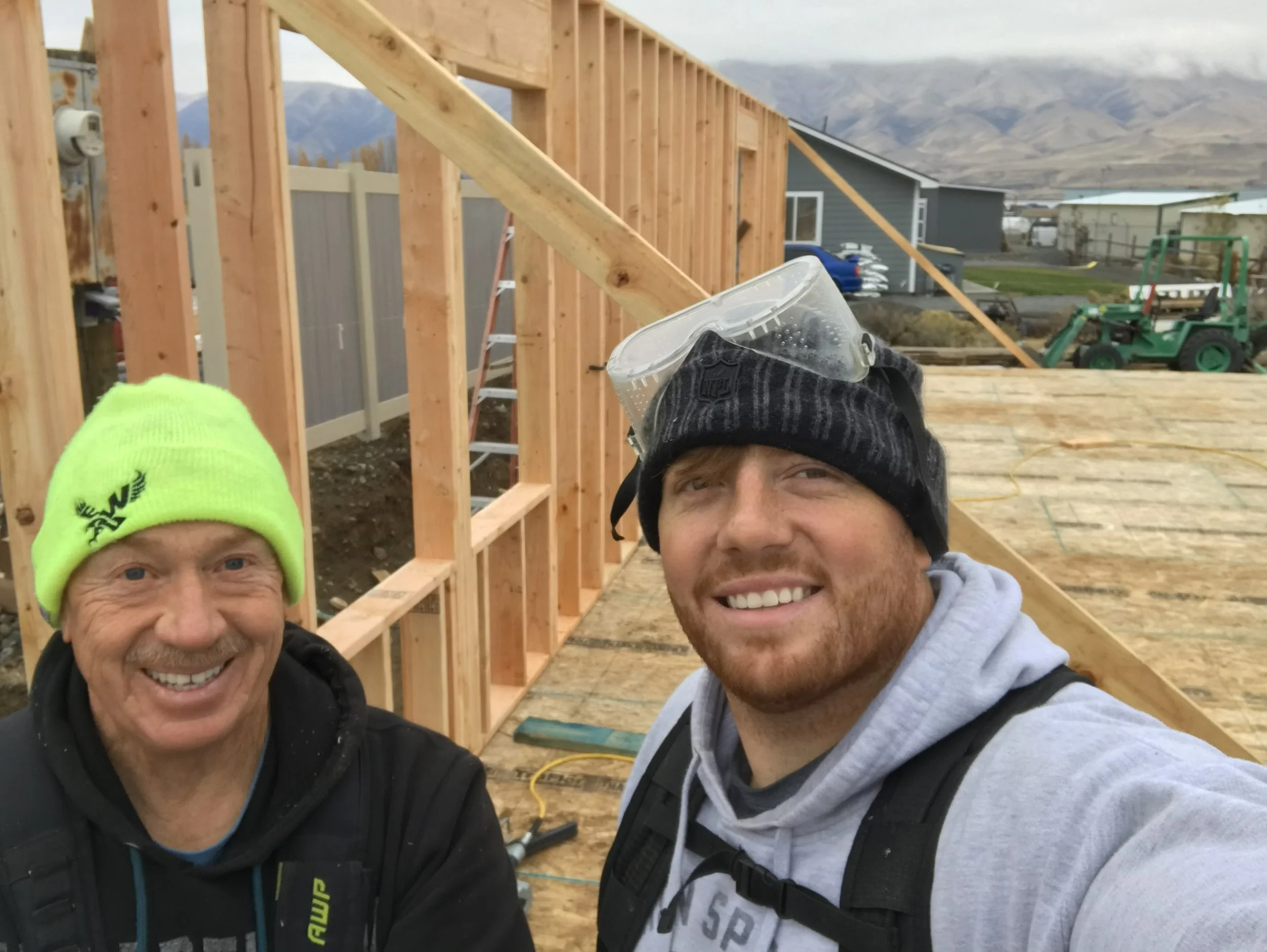 Two smiling men wearing warm clothes and beanies, taking a selfie at a house under construction with wooden framing, a ladder, and a scenic mountainous landscape in the background.