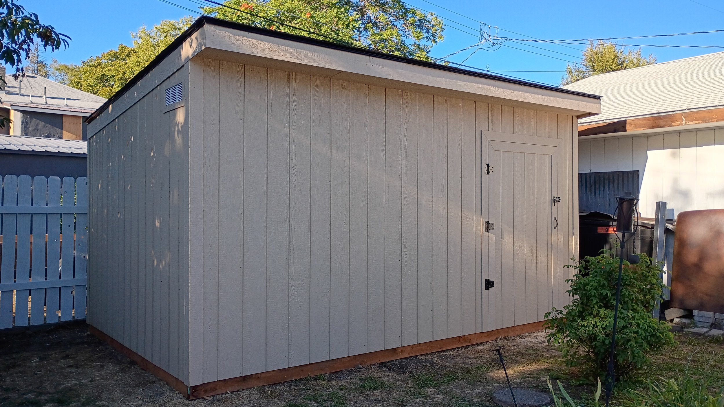 A small, beige wooden shed with a black roof in a backyard. There is a white fence on the left and various plants and objects around. A small bush is in front of the shed, and a garden ornament or tool is near the bush.