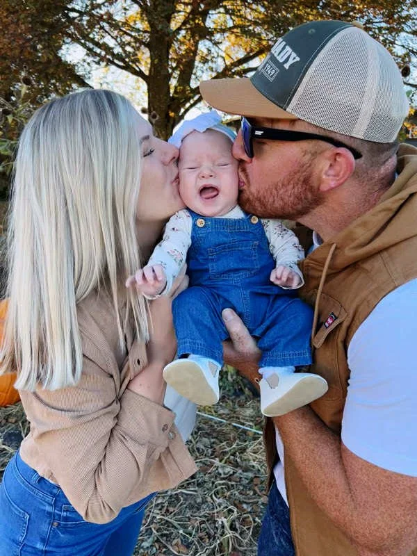A baby being kissed on each cheek by a woman and a man outdoors with trees in the background.