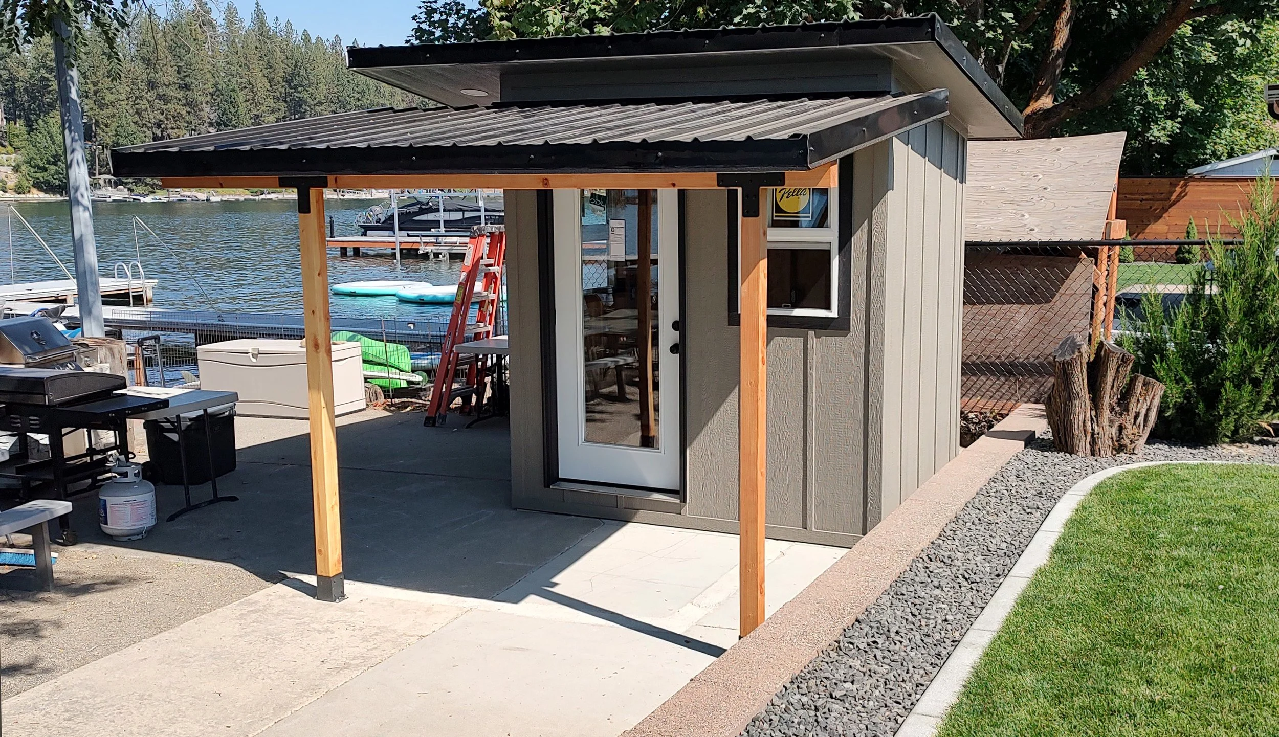Small outdoor shed by a lakeside with a white door, window, and partially built roof. Nearby outdoor furniture, a grill, and various boats and watercraft by the dock are visible in the background.