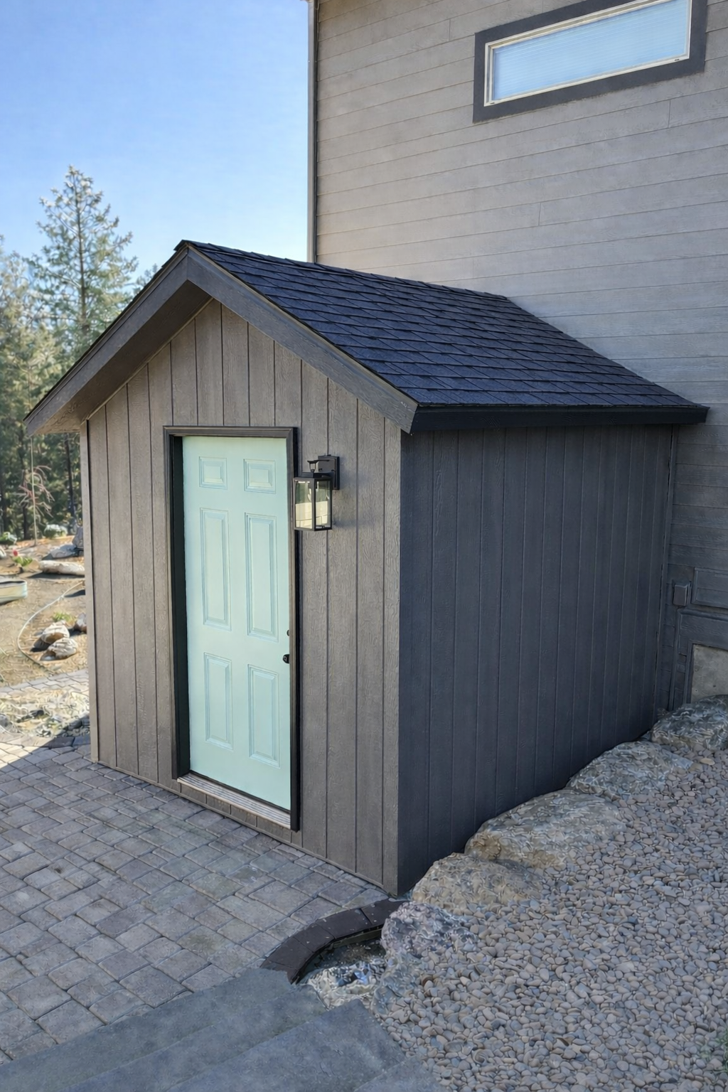 Small gray shed with a light green door, black trim, and an outdoor lantern on the wall, situated on a stone-paved pathway next to a larger house.